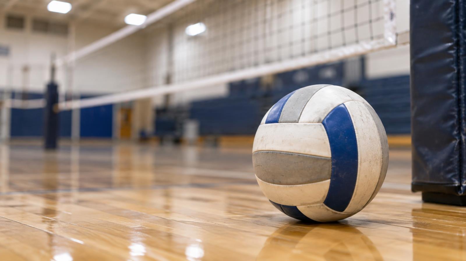 A volleyball sitting on a hardwood gym floor with a net in soft focus behind it.