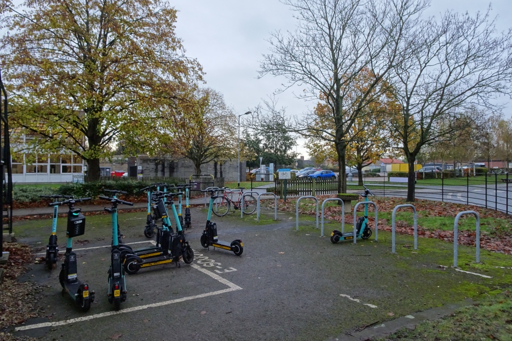 Several teal electric scooters parked near metal bike racks outside a building.