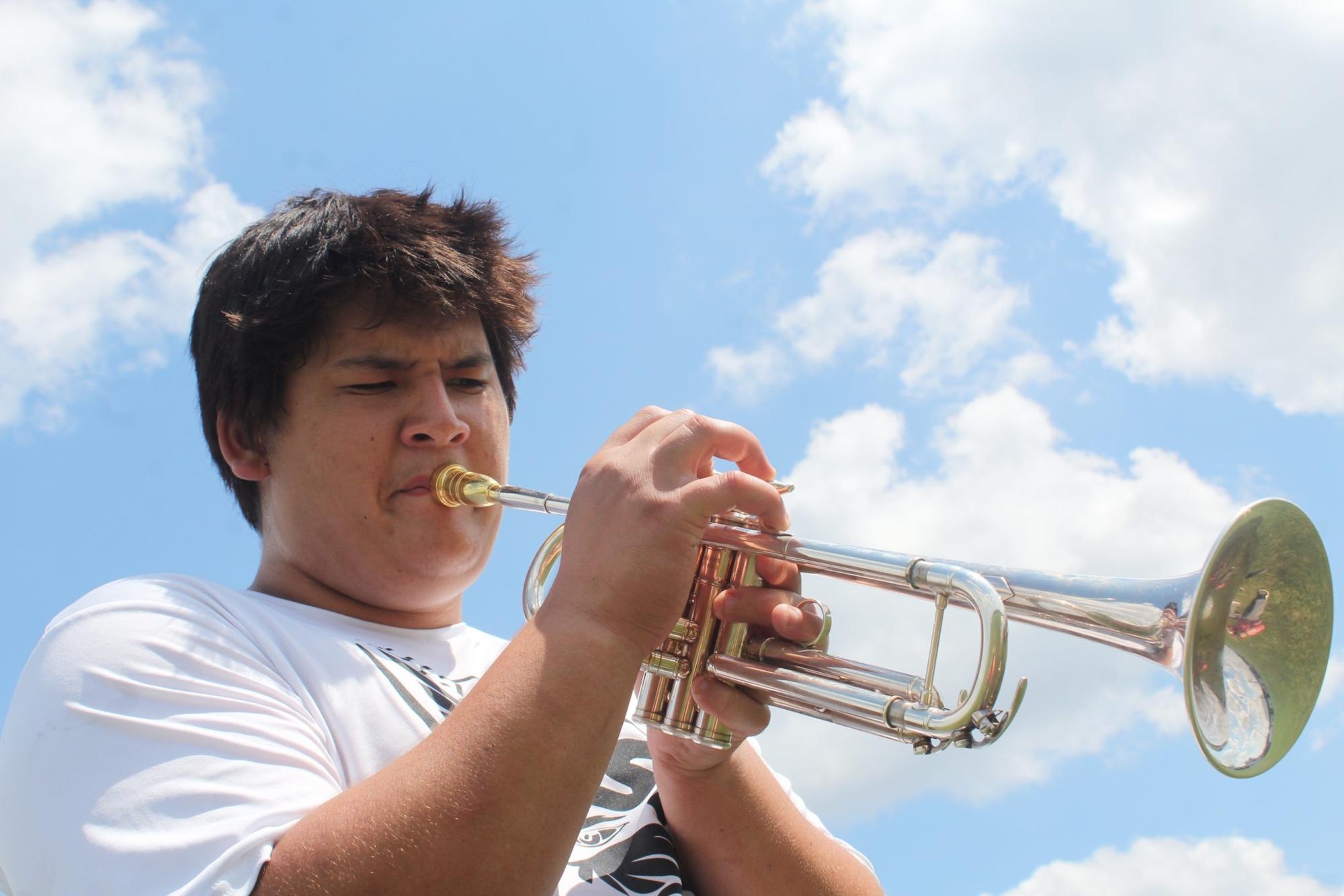 Young male student plays a silver trumpet outdoors against a partly cloudy sky.