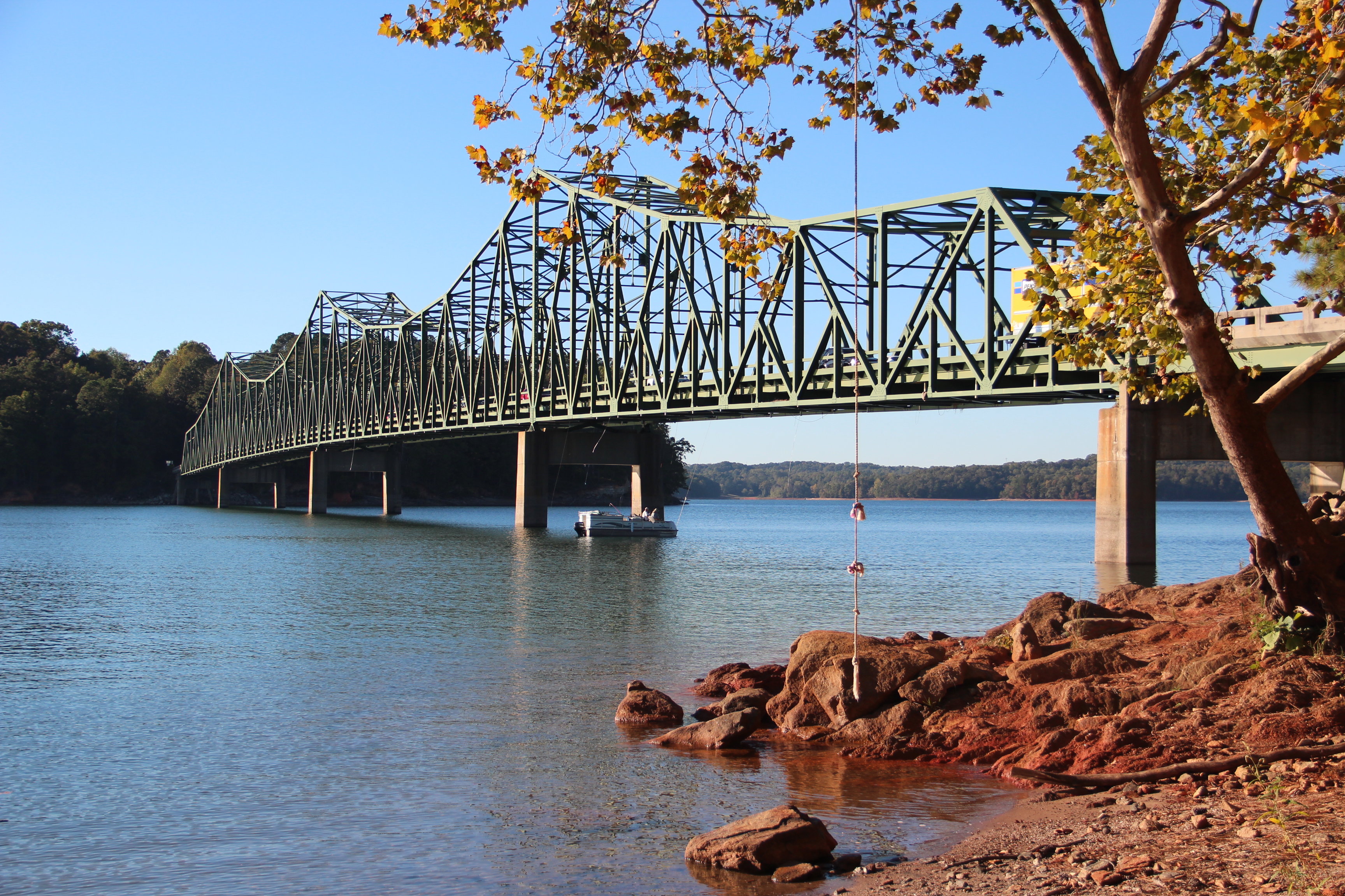 Steel truss bridge over calm blue lake water with red clay banks and autumn foliage.