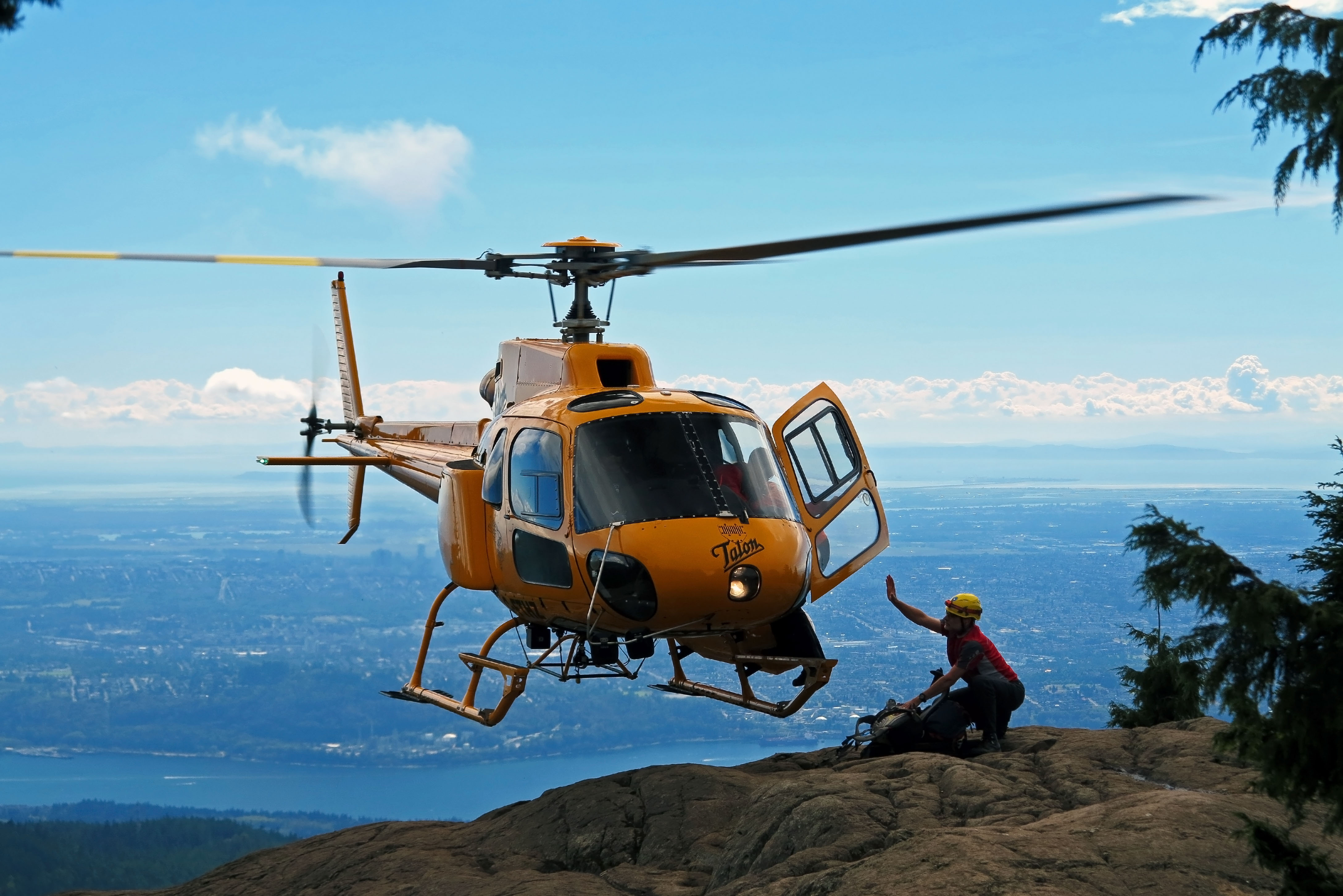 Yellow rescue helicopter hovering low over a rocky mountain ridge with a rescuer below.