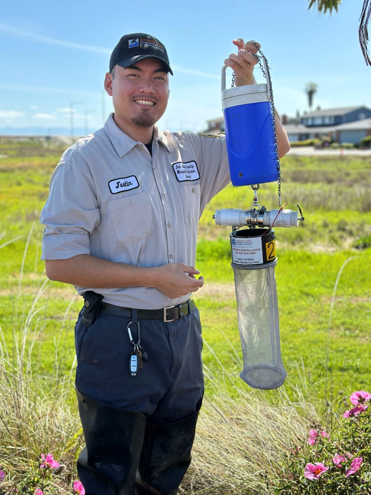 A uniformed technician holds a blue cylindrical mosquito trap with a mesh collection bag outdoors.