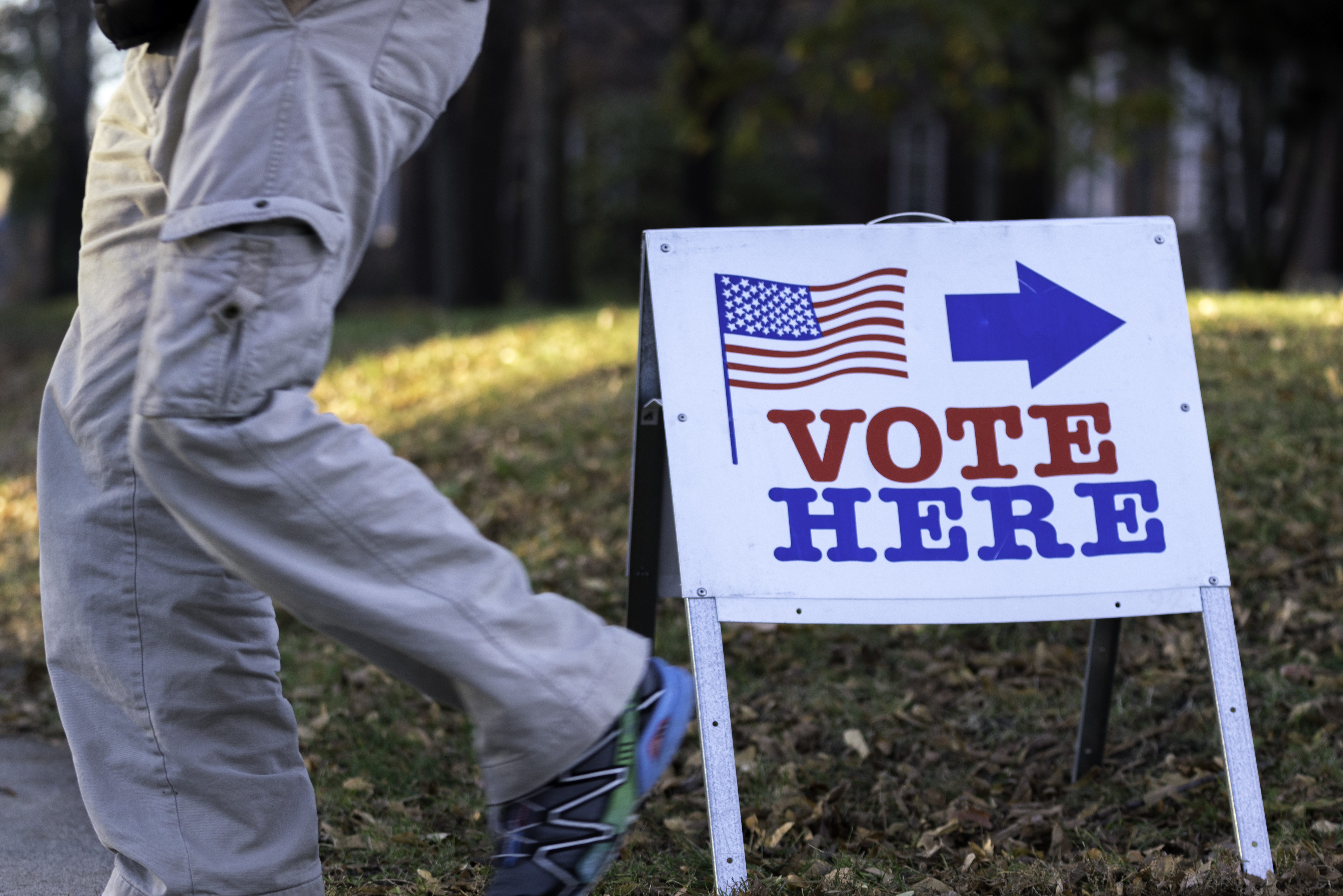 A portable 'Vote Here' sign with an American flag on a grassy area outside a building.