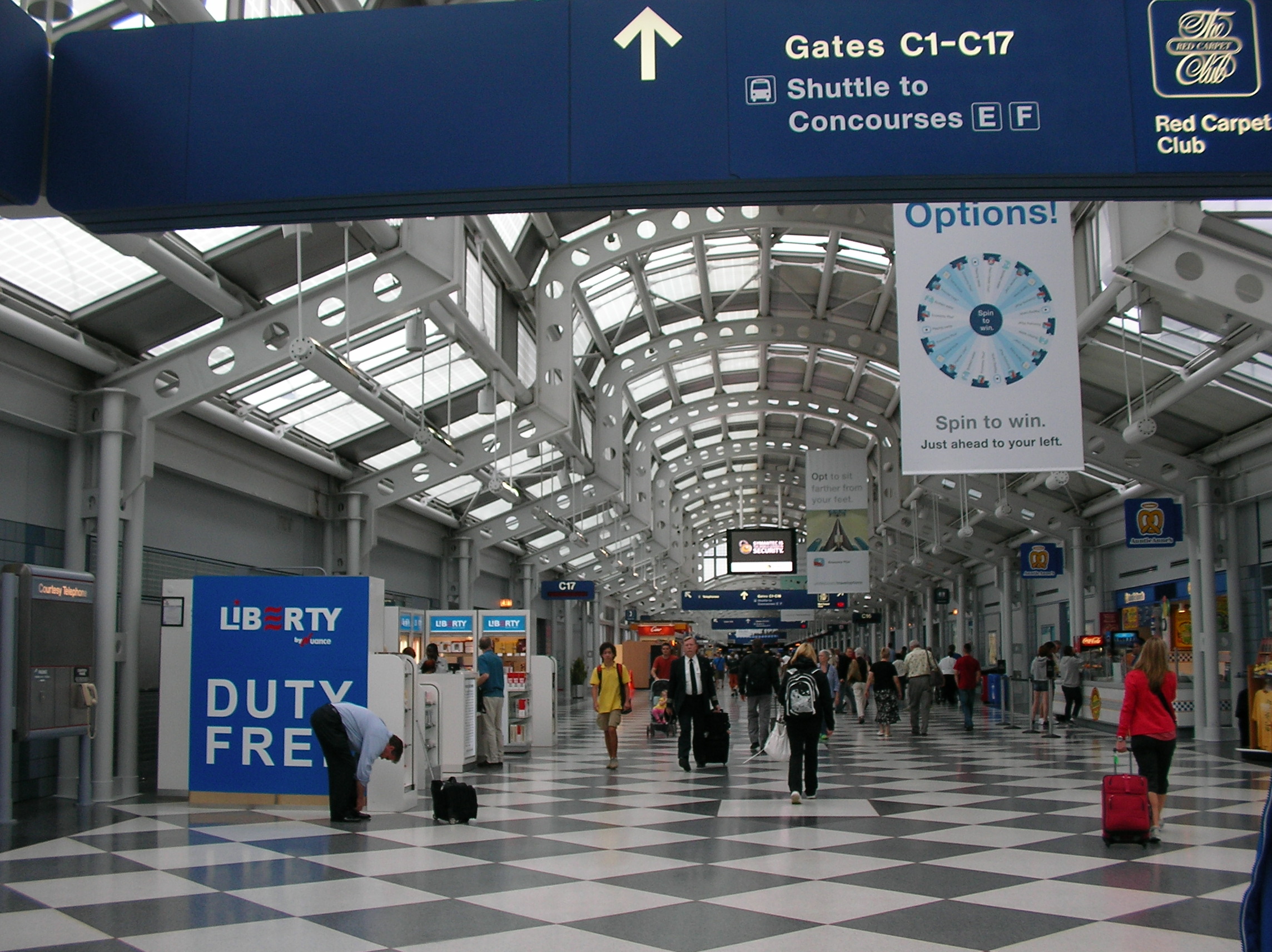 Wide airport concourse with travelers, gate signage, and an arched glass ceiling.