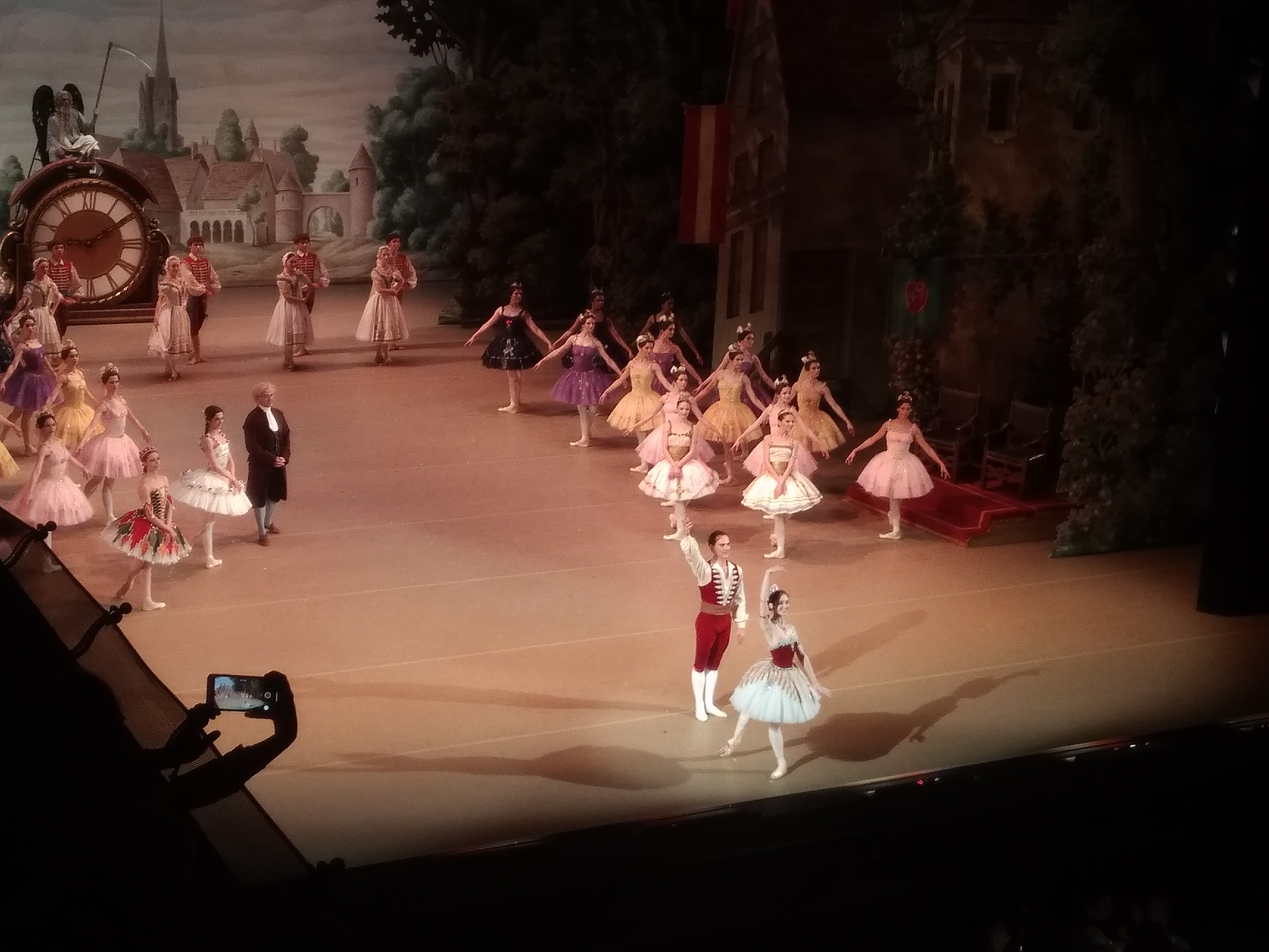 Ballet dancers in colorful tutus perform on a stage with a painted village backdrop and clock prop.