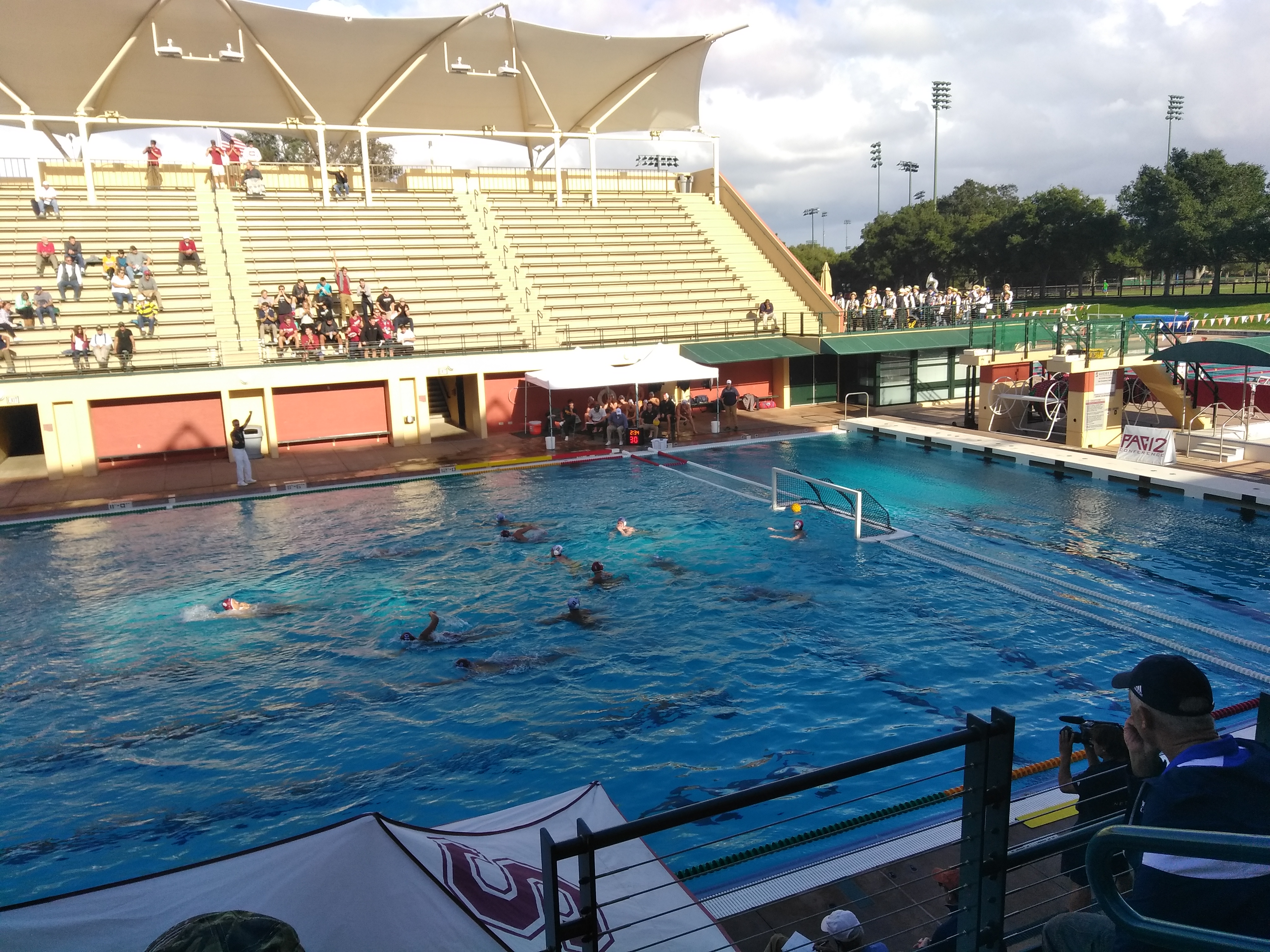 Water polo players in a pool during a match at an outdoor aquatic stadium with spectators in the stands.