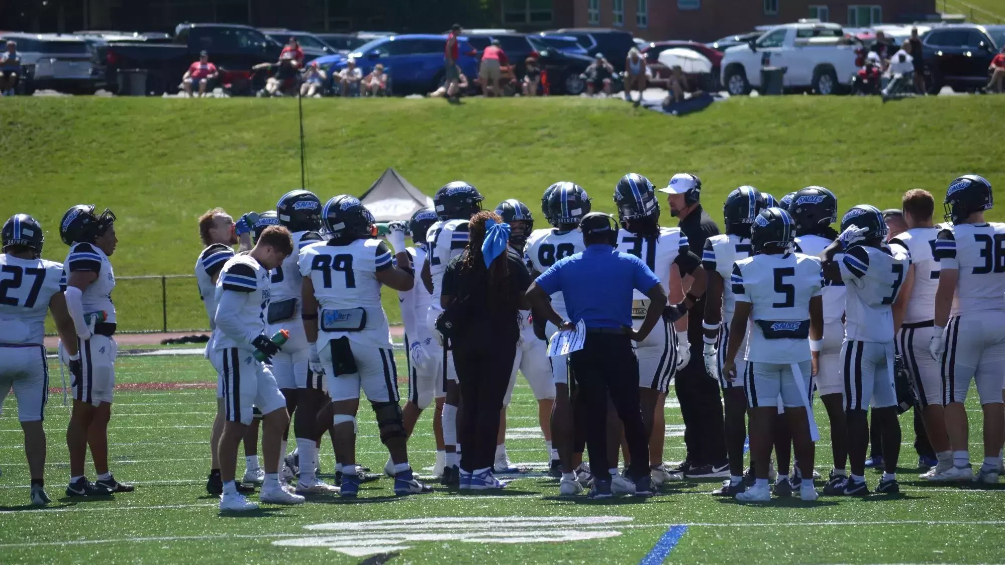 Football players in blue and white uniforms huddle on a turf field on a sunny day.