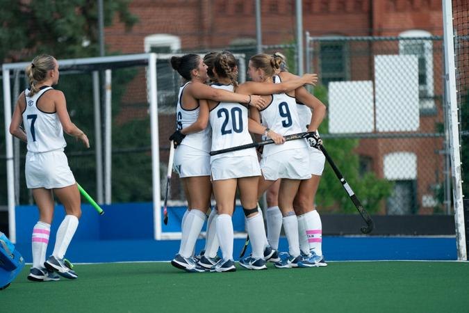 Female field hockey players in white uniforms celebrate together on a turf field.