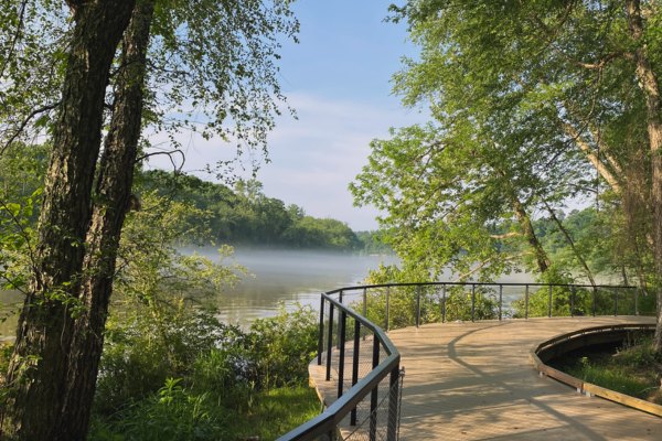 Curved wooden boardwalk with metal railings winding through green trees beside a misty river.