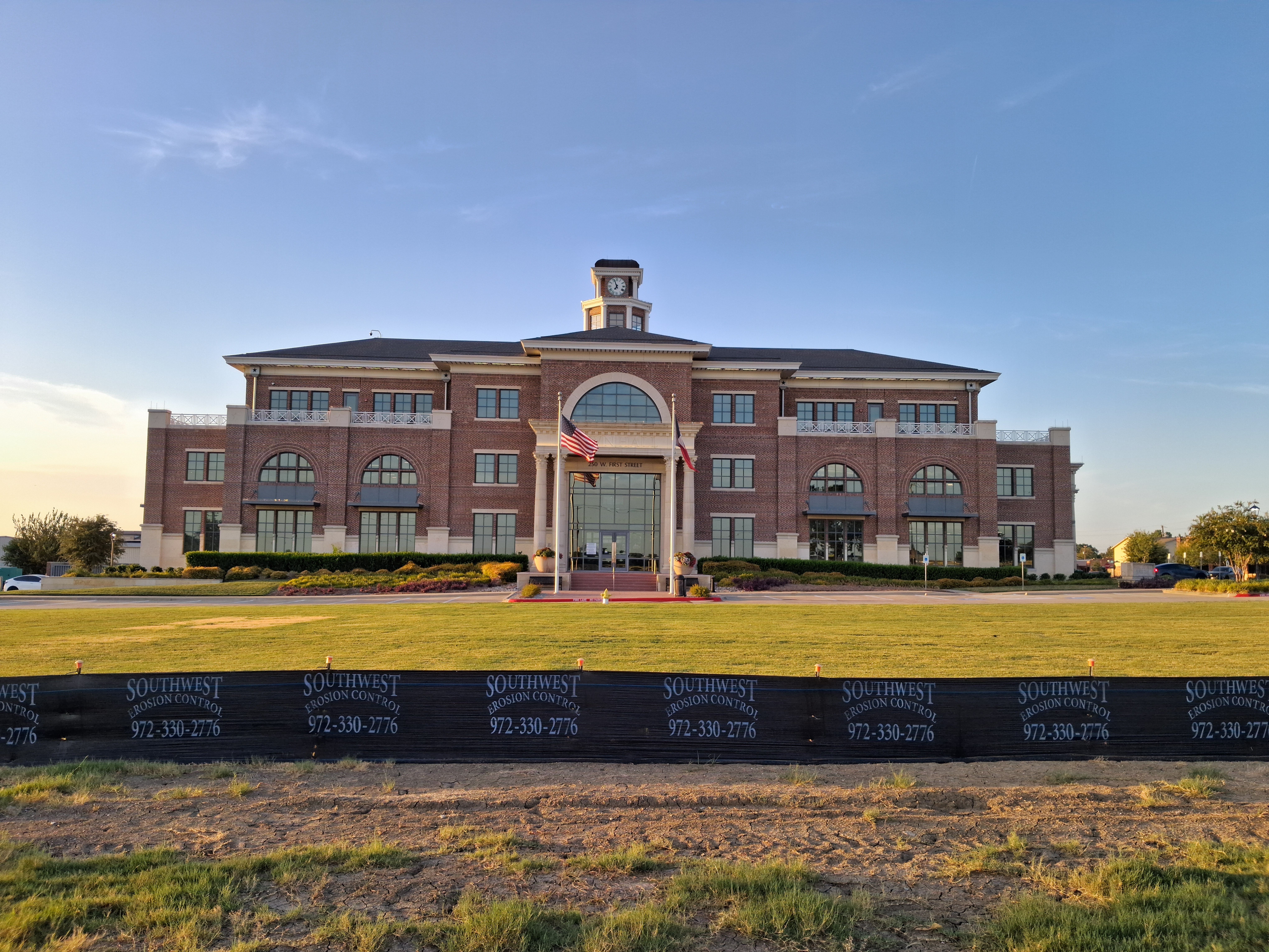 Front facade of a red-brick municipal building with a cupola and two flags.