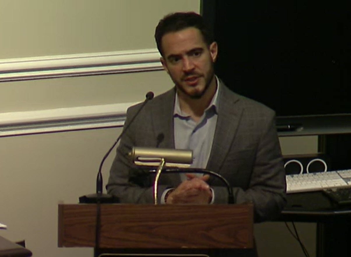A man in a gray blazer stands at a wooden podium with a microphone in a meeting room.