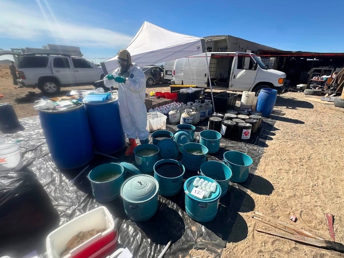 Person in white hazmat suit standing among large blue drums and chemical containers in a desert setting.