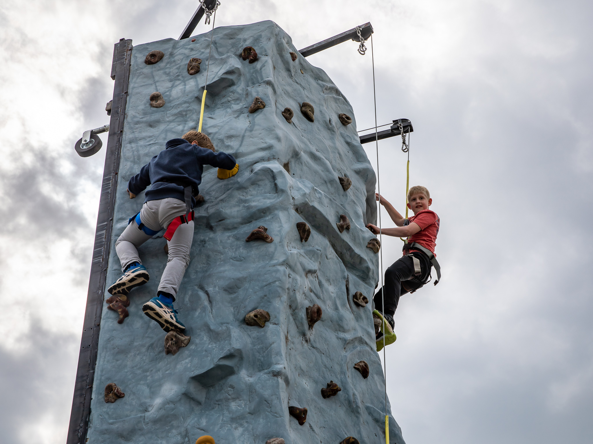 Two children in safety harnesses climb a colorful portable outdoor rock wall.