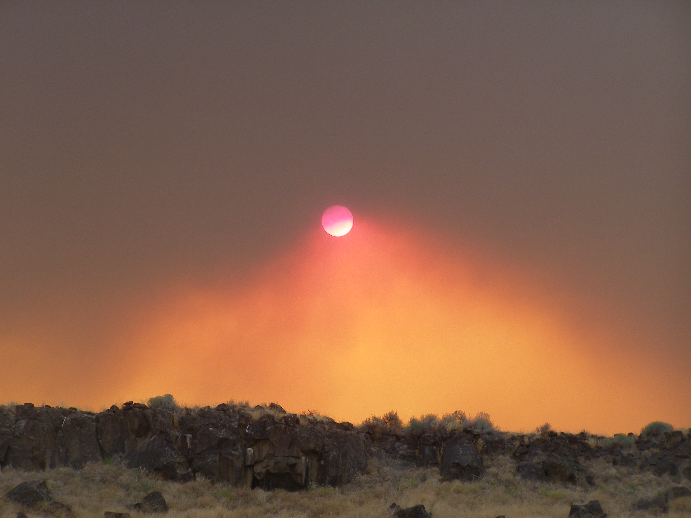 Massive smoke plume from a wildfire turns the sky orange over a rocky desert landscape.