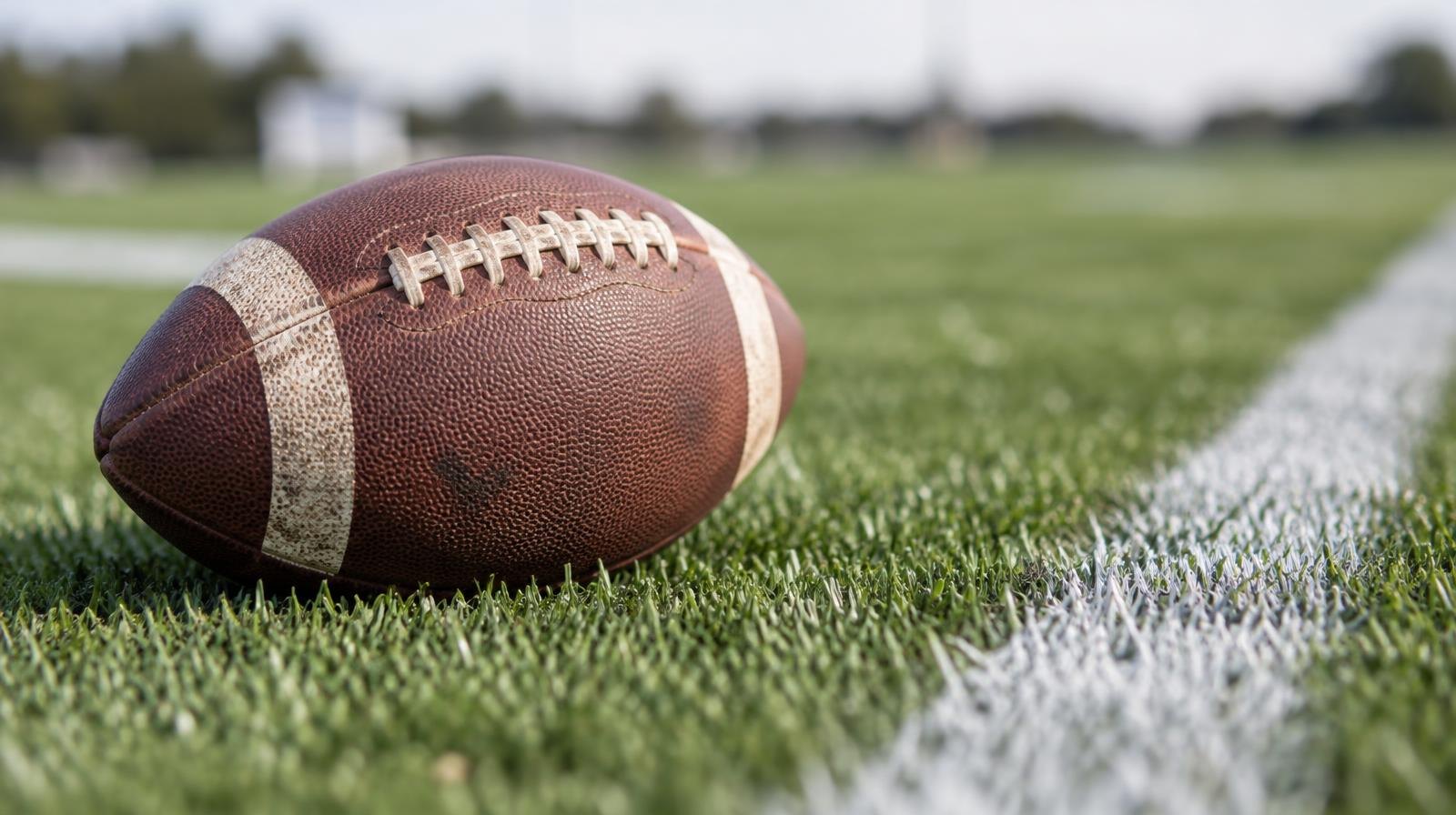 An American football resting on green grass next to a white yard line.