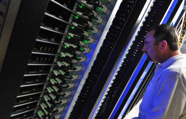 A man examining rows of server racks with green indicator lights in a blue-lit data center aisle.