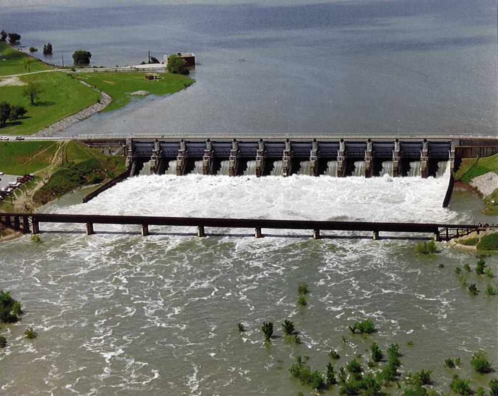 Aerial view of a dam with multiple open spillway gates releasing turbulent white water.