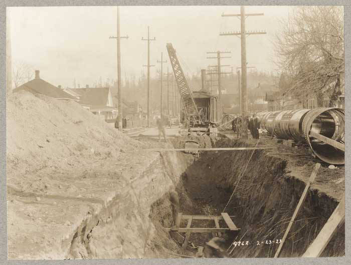 Workers and a crane lower a large steel pipe into a street trench.