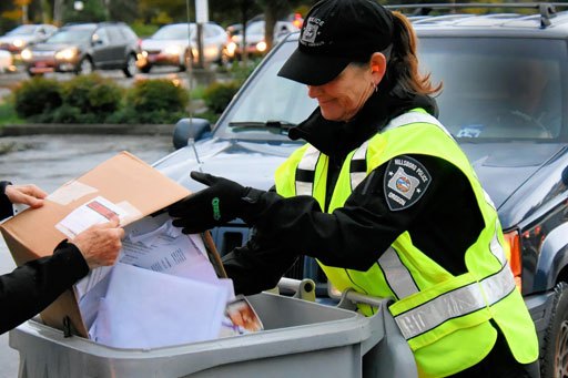 A police officer in a yellow vest directs a person depositing boxes into a gray collection bin.