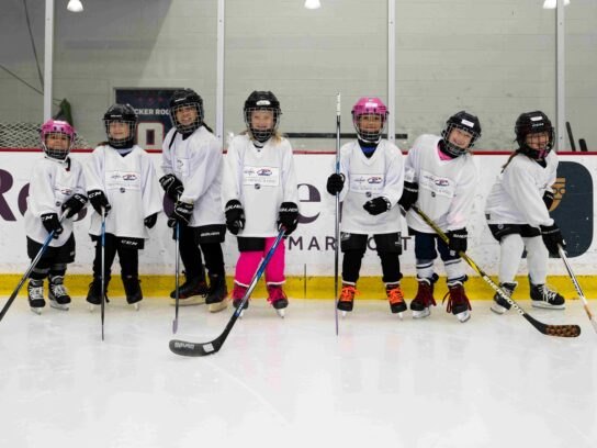 Six young girls in white hockey jerseys and helmets standing in a line on an ice rink holding hockey sticks.