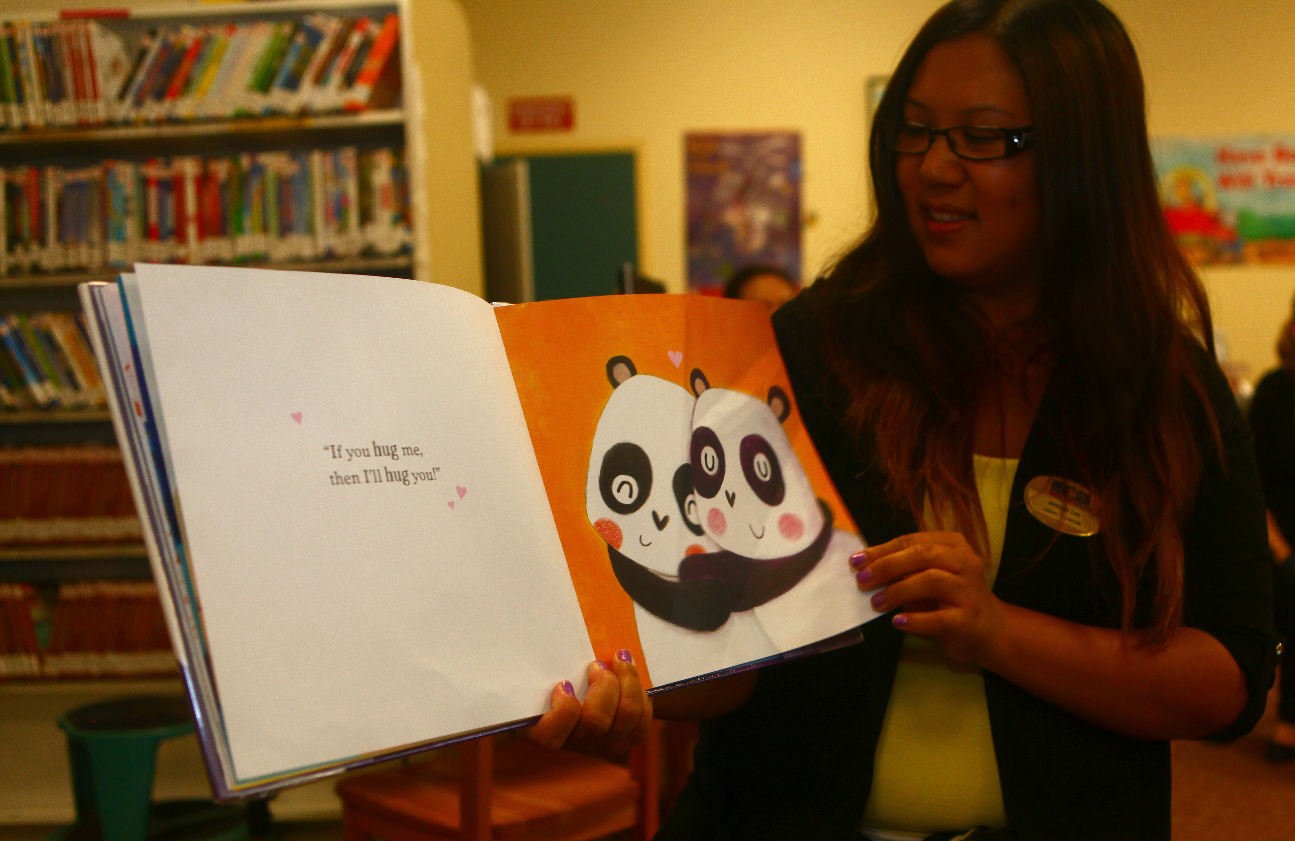 A woman holds open a colorful children's picture book in a library with bookshelves behind her.