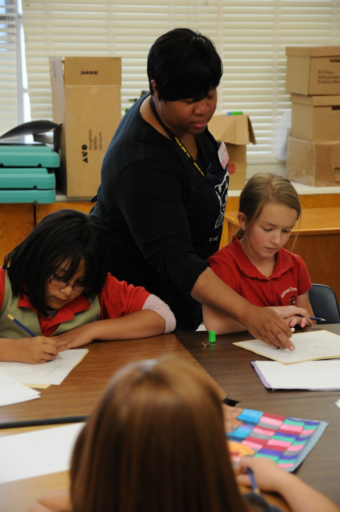 An adult woman leans over a table helping two young girls with written work in a classroom.