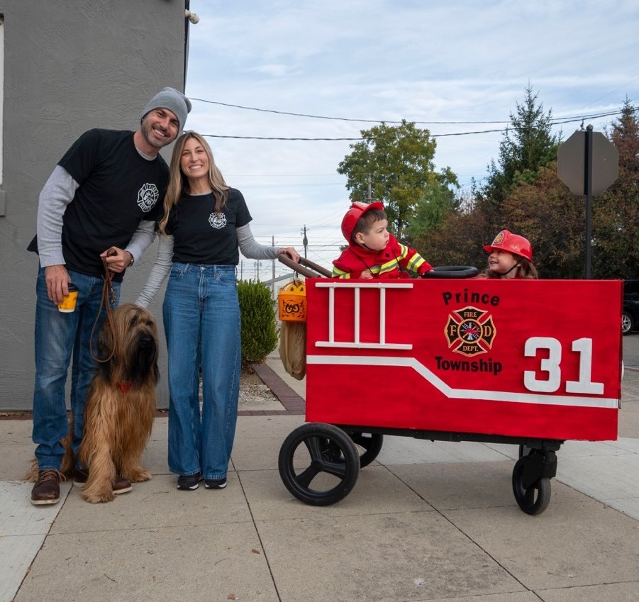 Two small children in red firefighter costumes sitting in a red wagon decorated as a fire truck on a sidewalk.