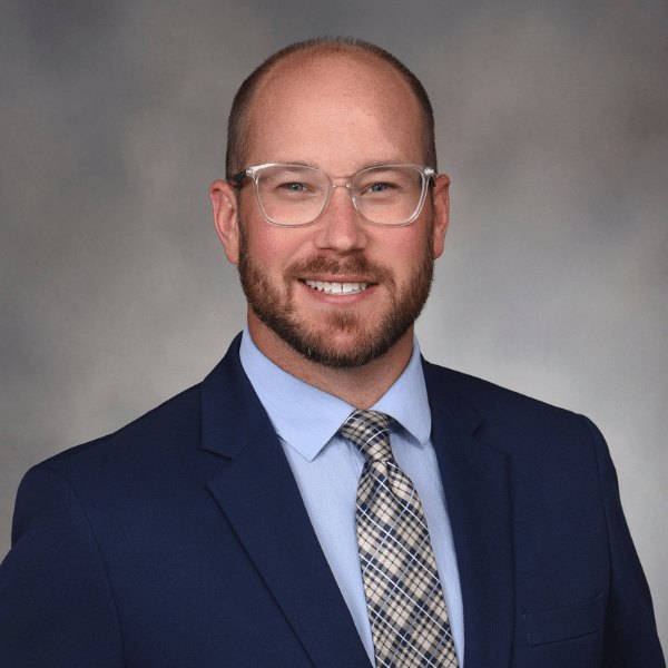Professional headshot of a bearded man in a navy blazer and plaid tie.