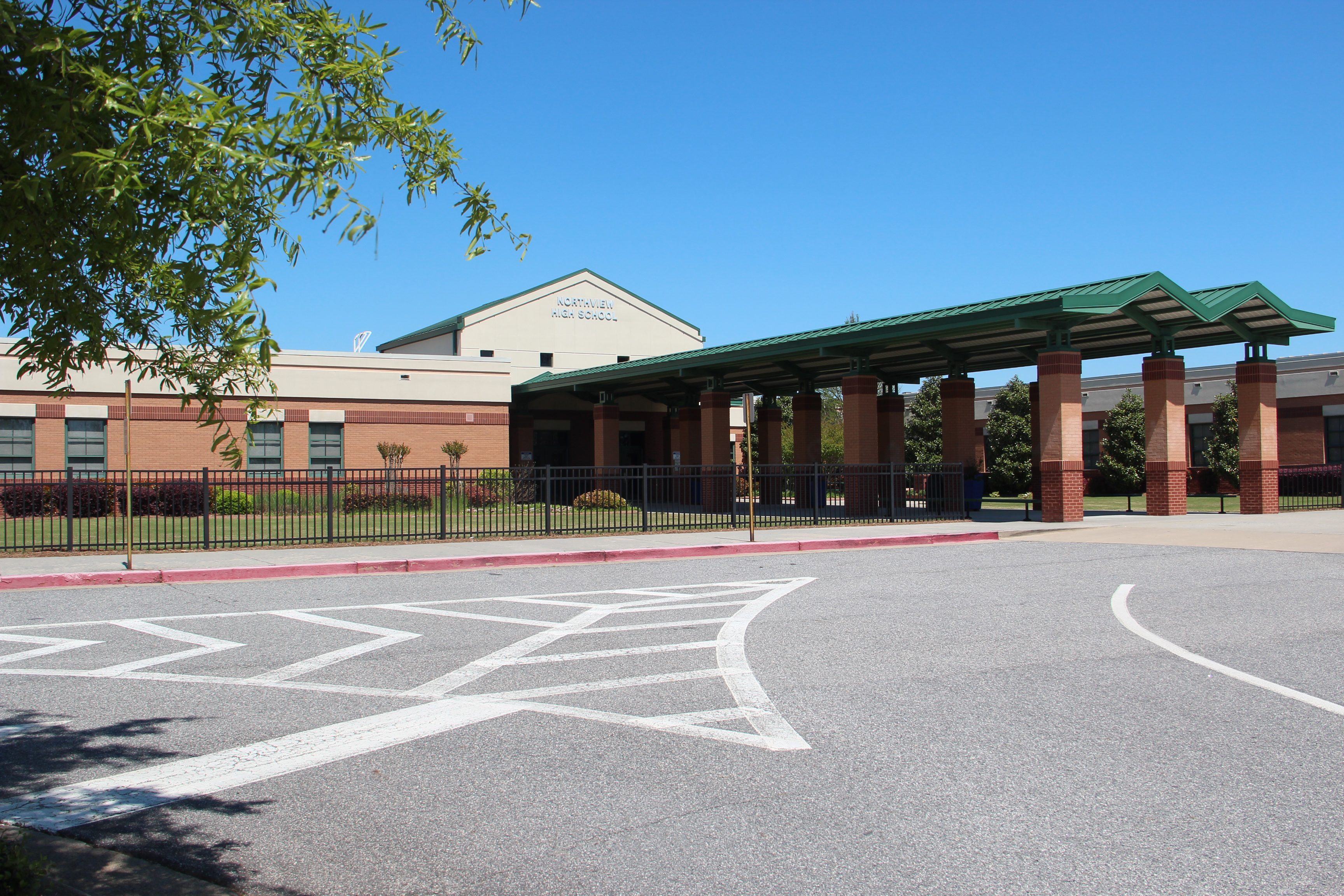Brick school building with green-roofed entrance canopy and iron fencing on a sunny day.