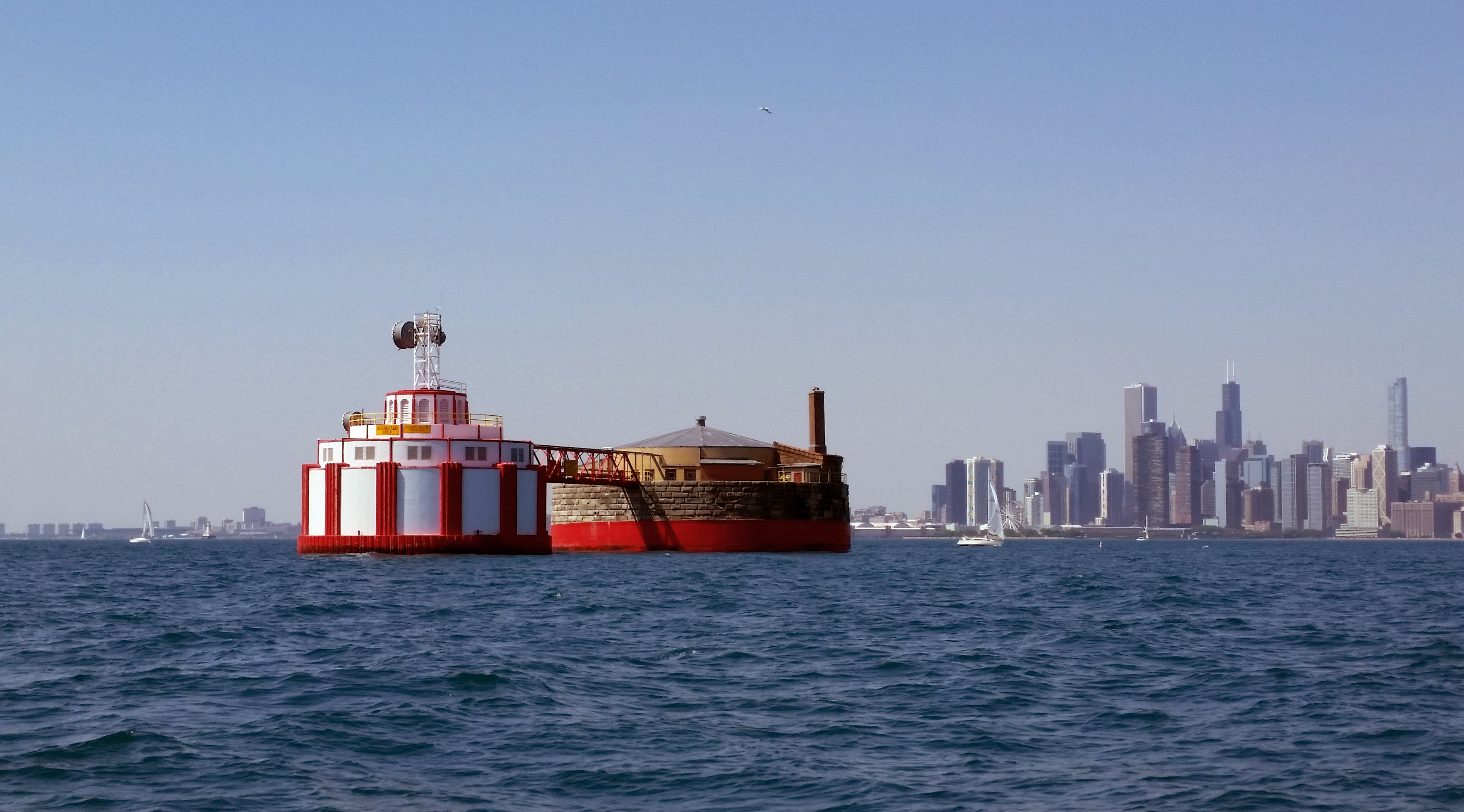 Two red-and-white water intake cribs in Lake Michigan, connected by a footbridge, with the Chicago skyline in the background.