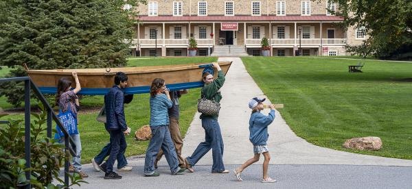 A group of people carry a wooden flat-bottomed boat across a campus lawn.