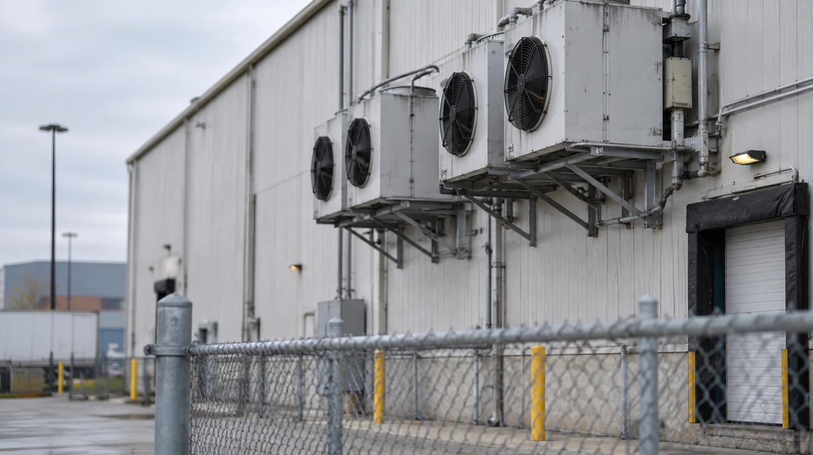Large industrial warehouse exterior with wall-mounted refrigeration units and a chain-link fence.