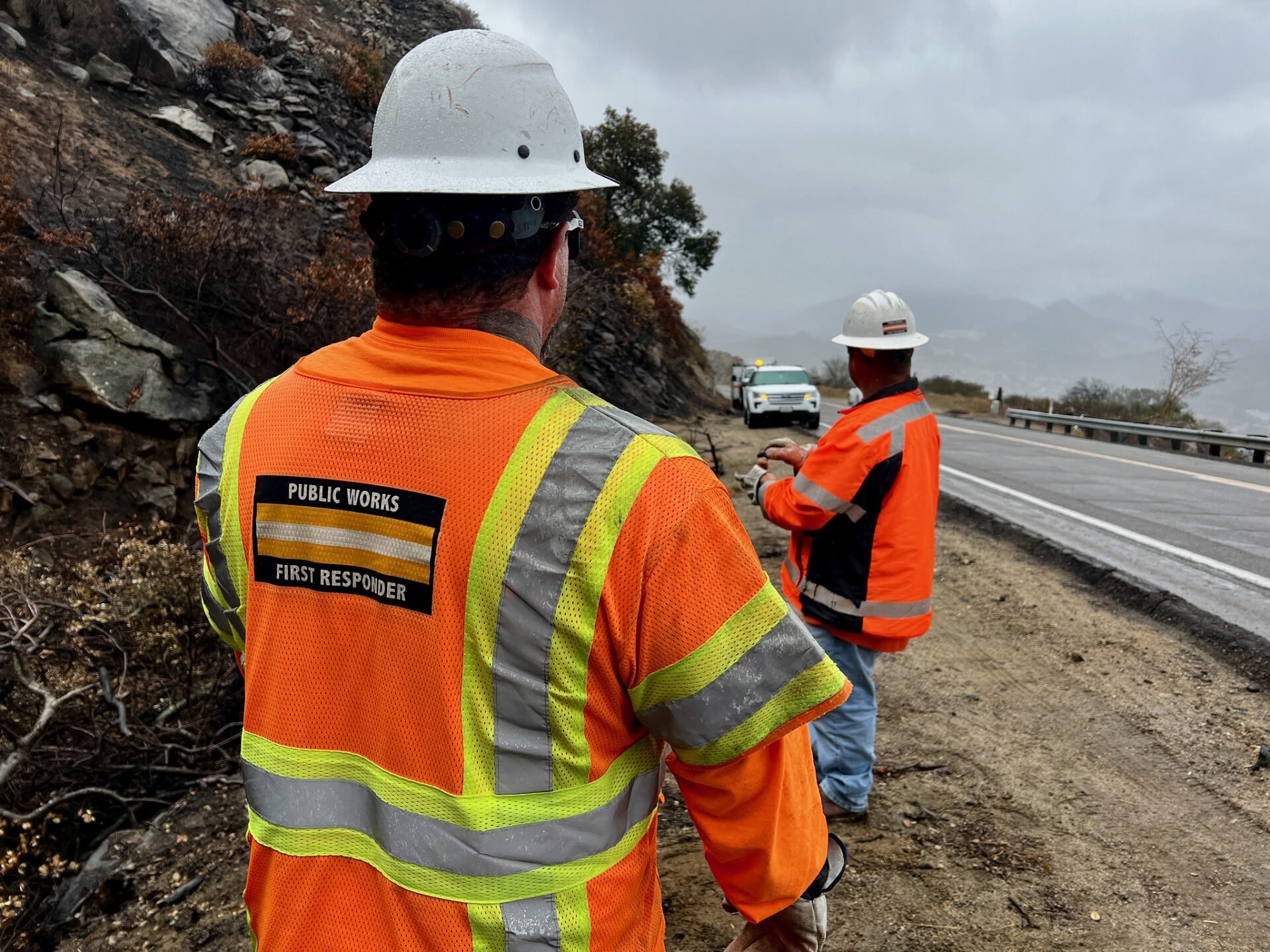 Two workers in orange safety vests and hard hats stand on a wet road shoulder beside a rocky hillside.