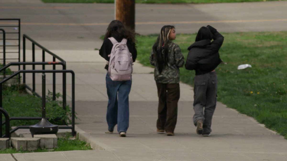 Three young people with backpacks walking along a sidewalk, photographed from behind.