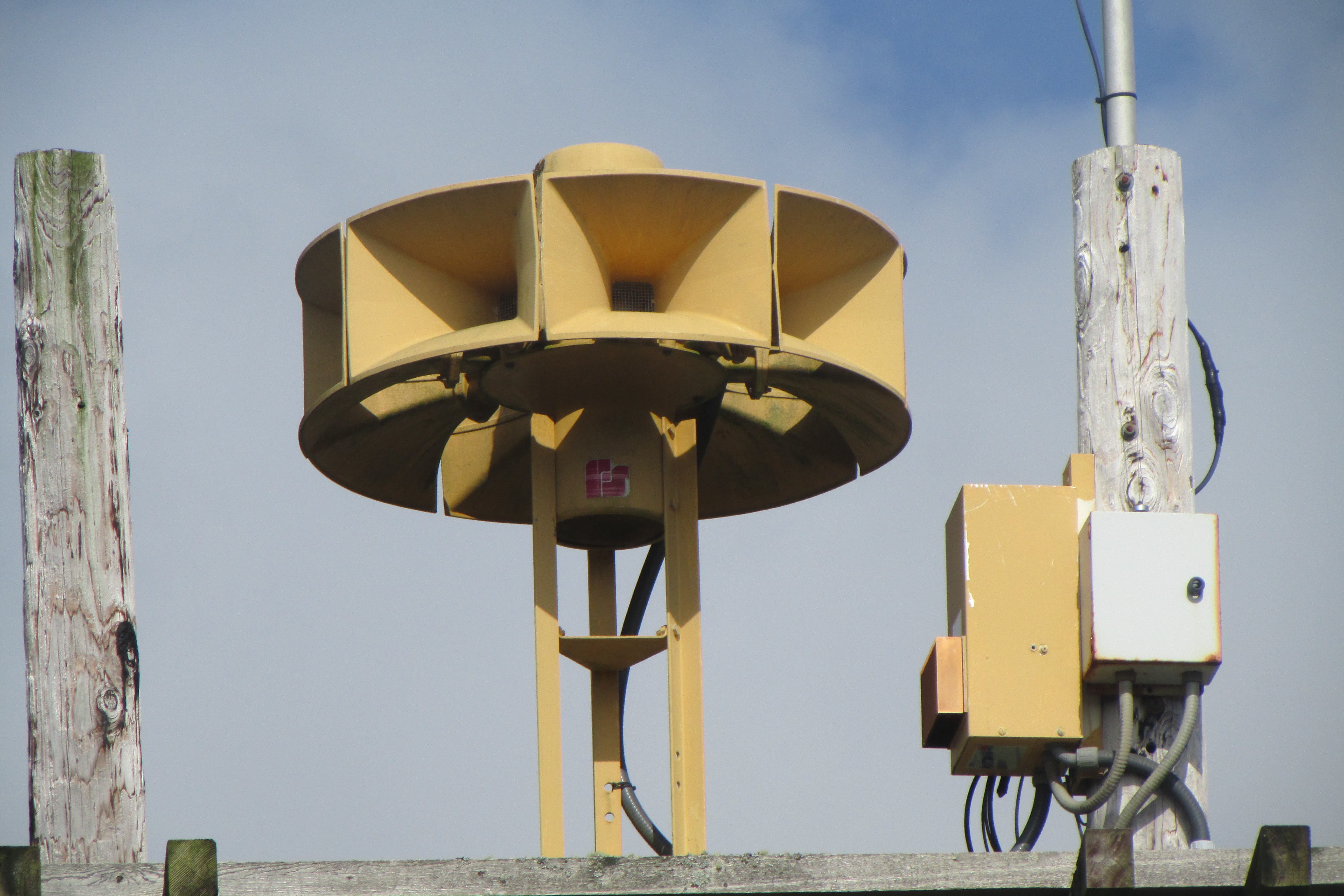 A yellow multi-directional outdoor warning siren on a metal pole against a partly cloudy sky.