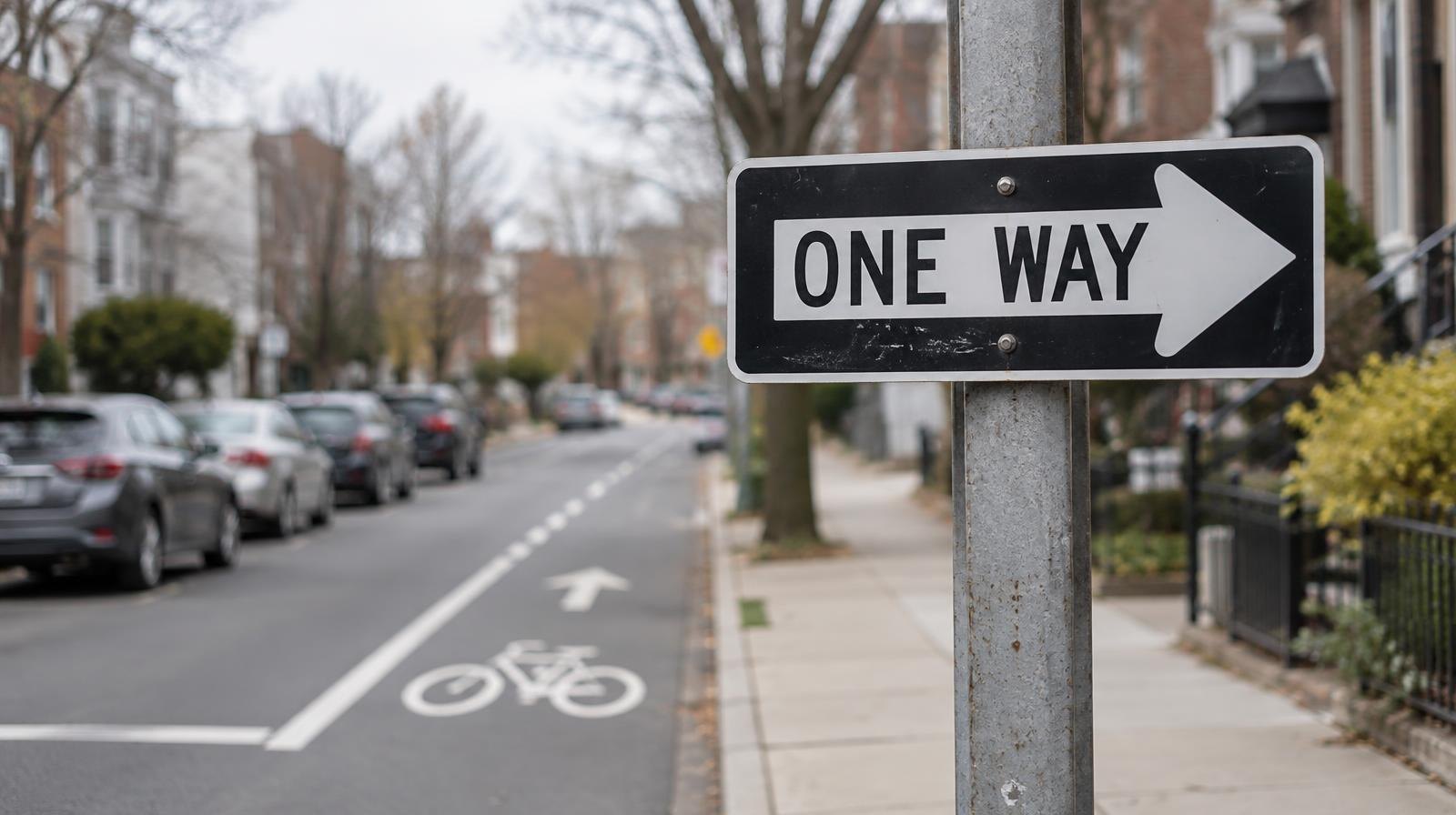 One-way street sign at an urban intersection with a painted bike lane alongside parked cars.