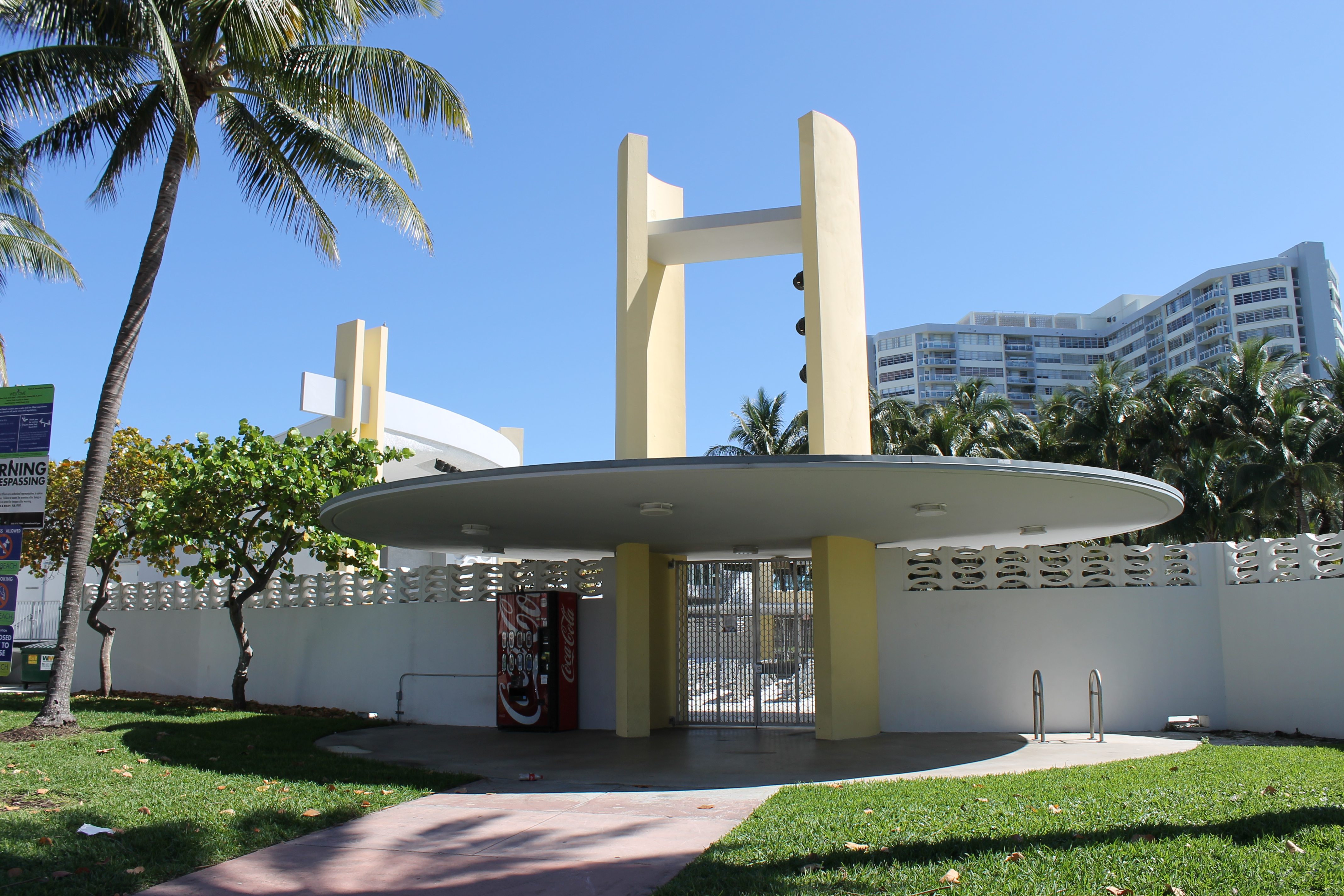 Mid-century modern park pavilion with oval canopy roof, yellow columns, and palm trees.