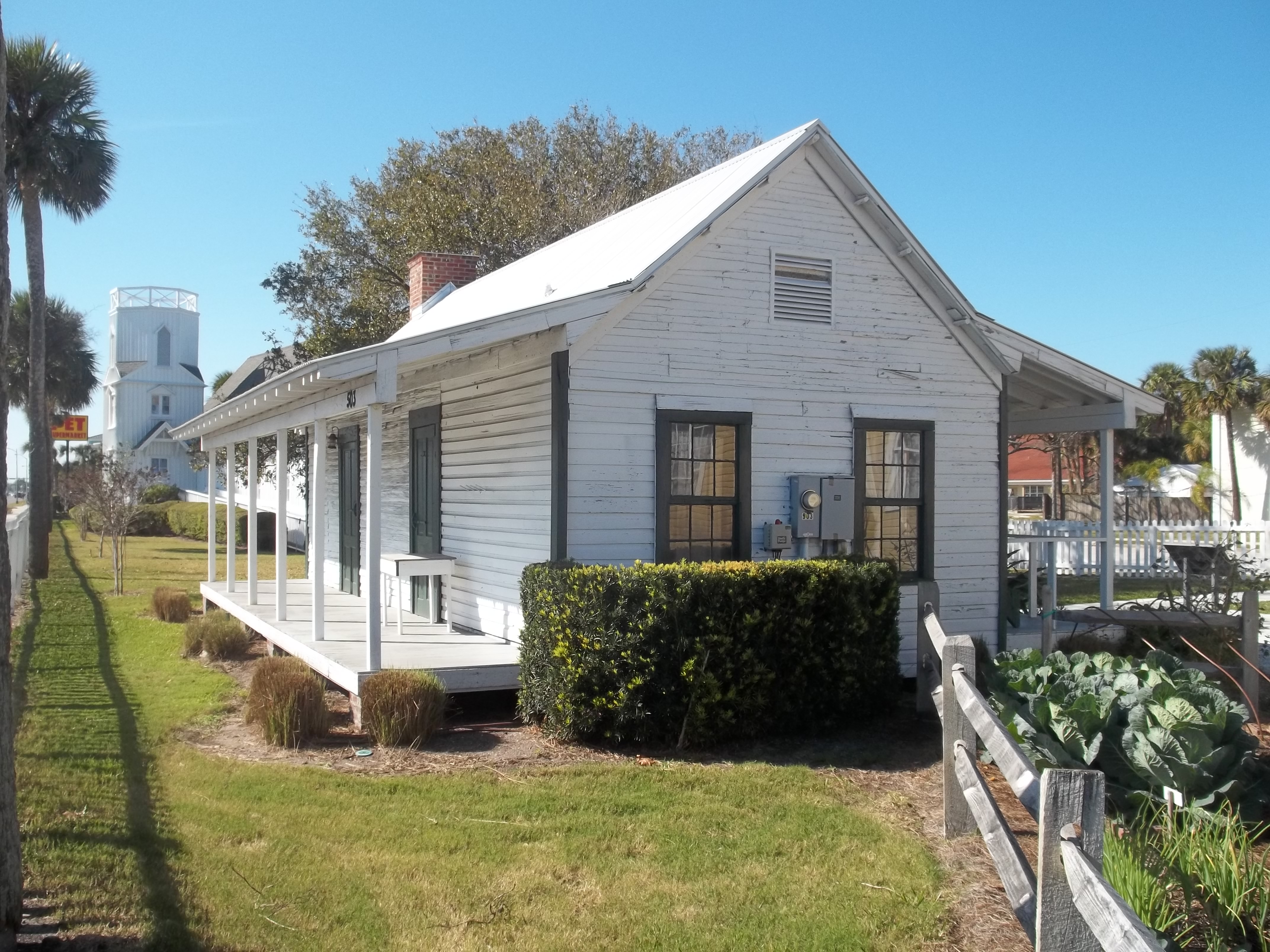 Small white historic cottage with front porch, garden, and palm trees on a sunny day.