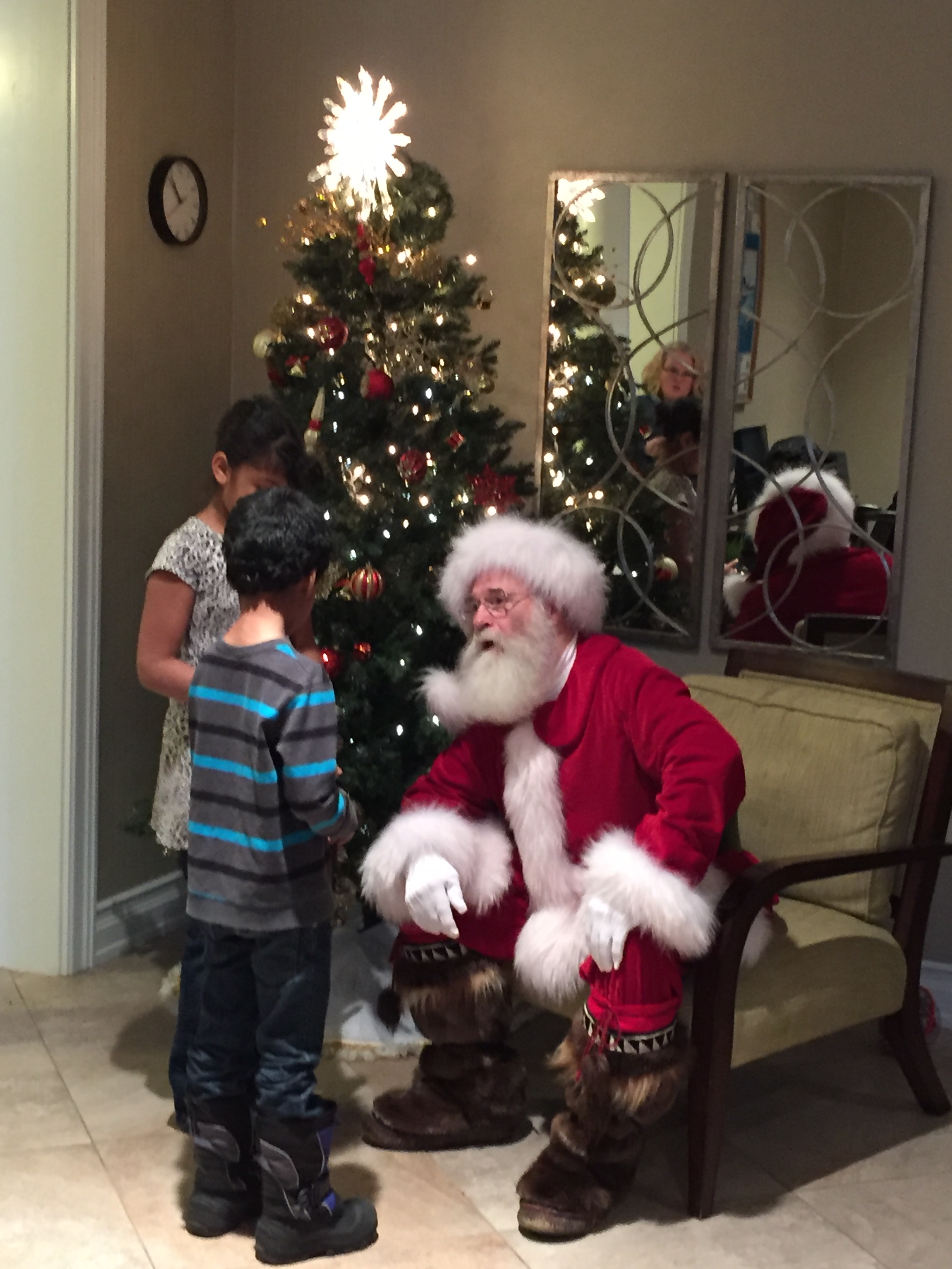 Santa Claus seated and speaking with two young children near a lit Christmas tree.