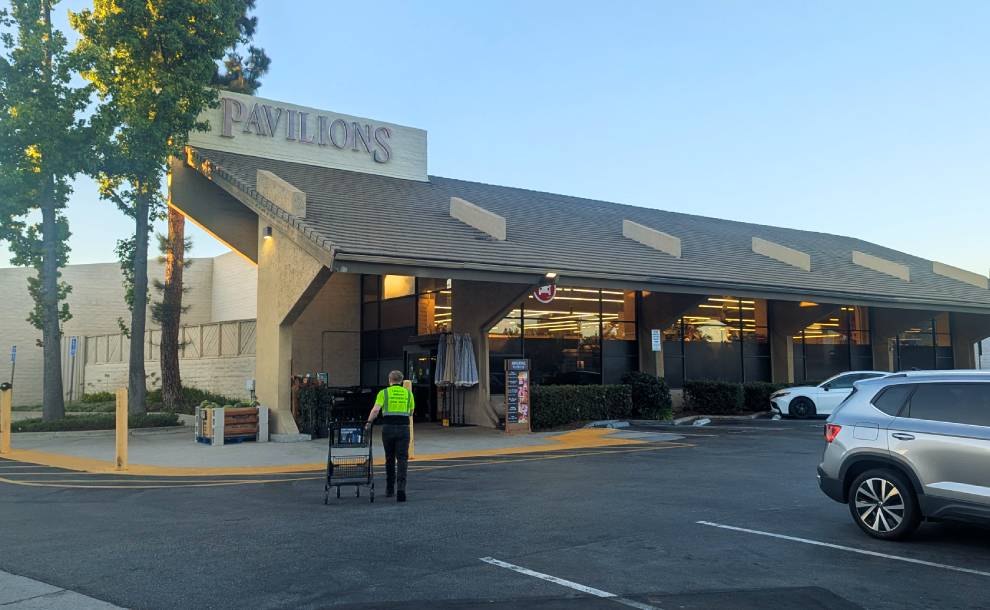 Exterior of a Pavilions supermarket with its A-frame roof, signage, parking lot, and a person pushing a shopping cart toward the entrance.