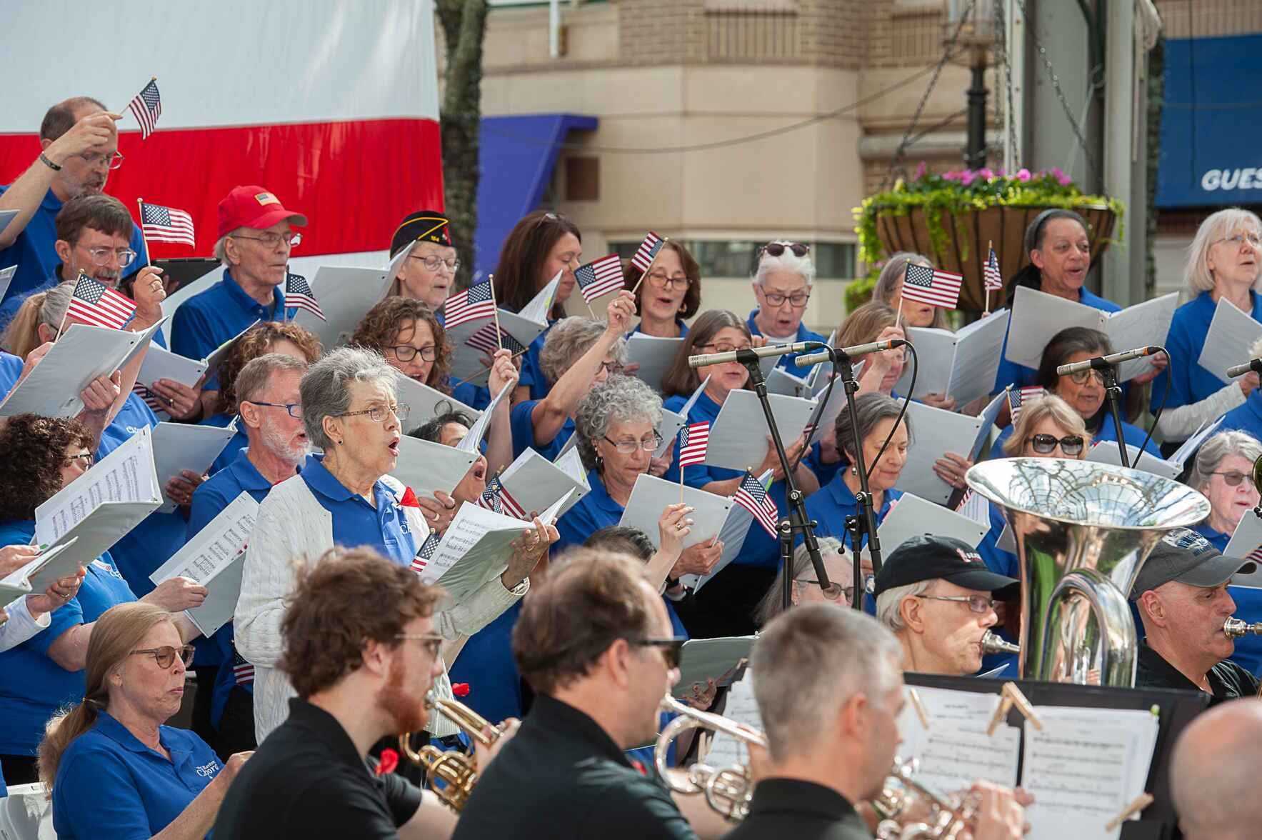 Community chorus and orchestra performing outdoors, members in blue shirts holding sheet music and small American flags.