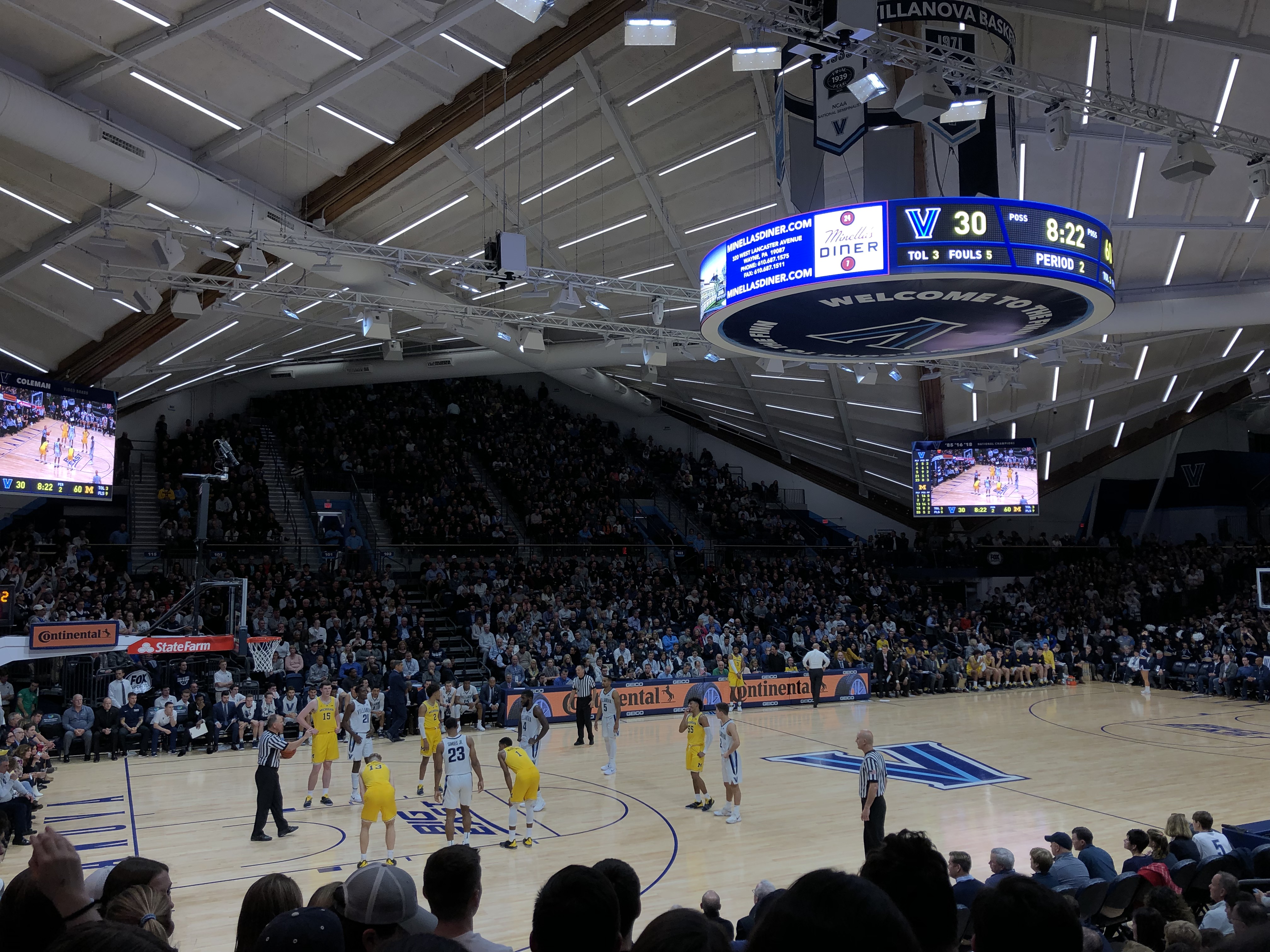 Interior of a college basketball arena with a packed crowd and players on the court.