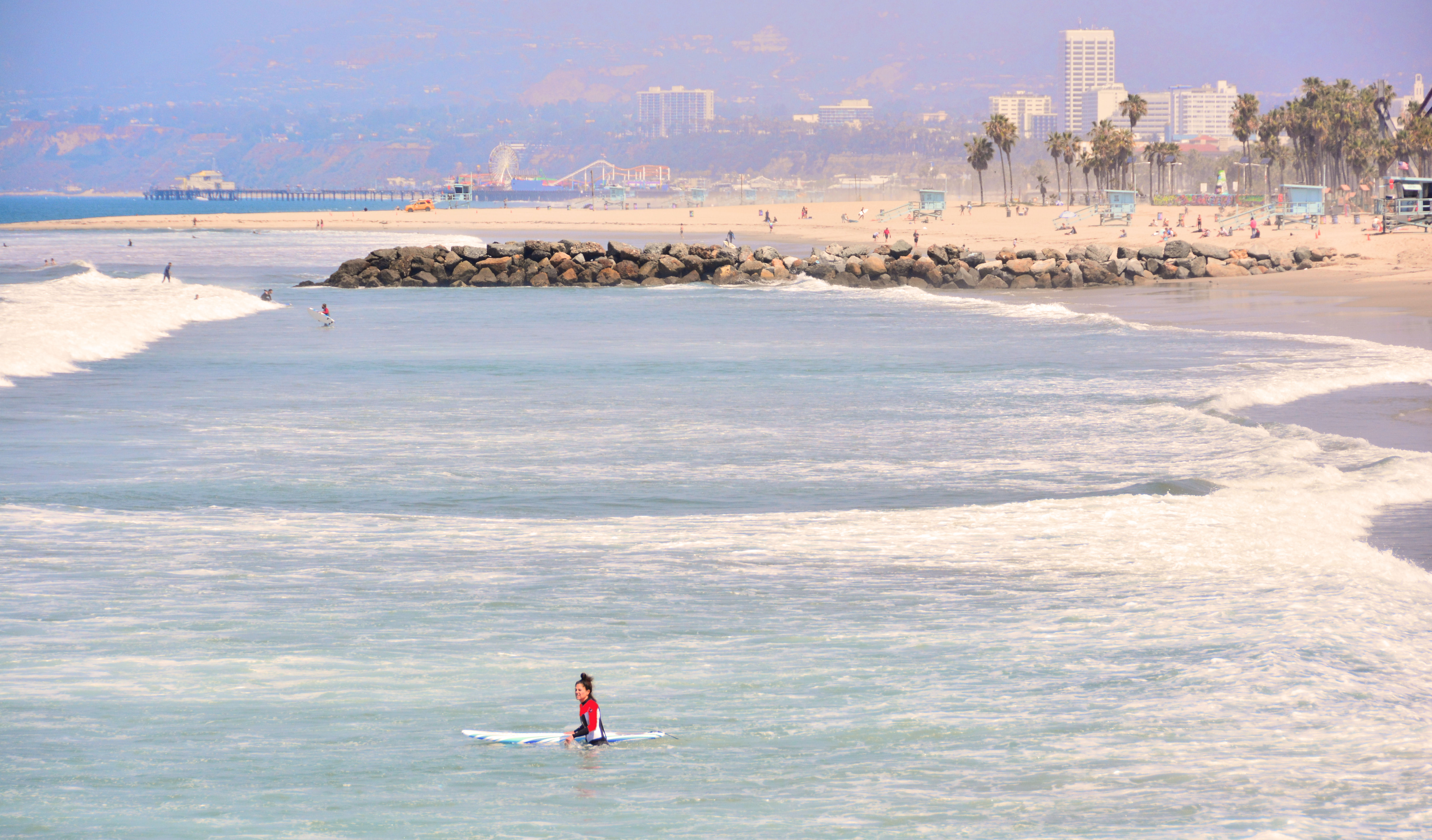 Surfer in shallow water near a rock jetty with Santa Monica Pier visible in the background.