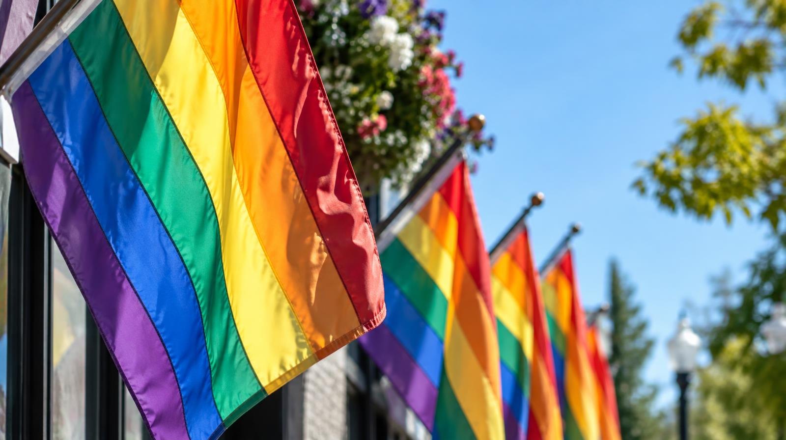 A row of colorful rainbow pride flags hanging outdoors against a blue sky.