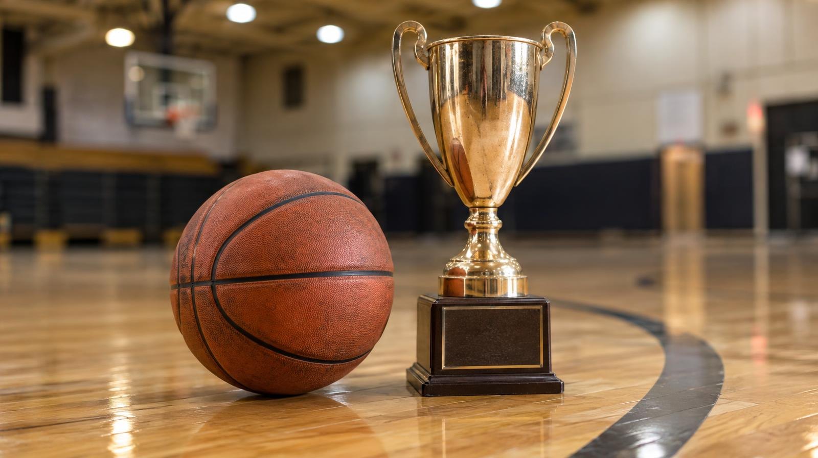 A basketball on a hardwood gym floor near a three-point arc with a trophy beside it.
