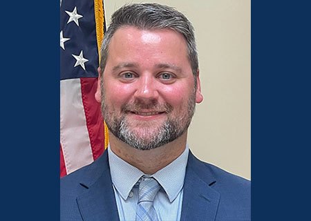 Professional headshot of a man in a blue suit and patterned tie in front of an American flag.