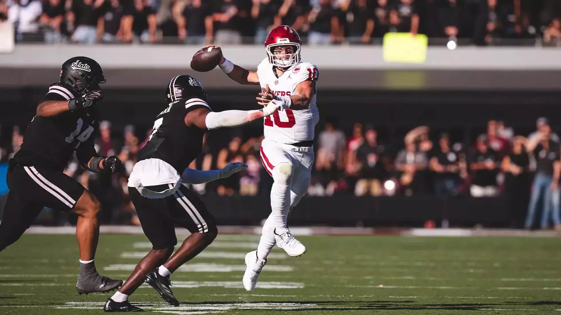 Quarterback in white uniform and red helmet throws a pass under pressure from defenders.