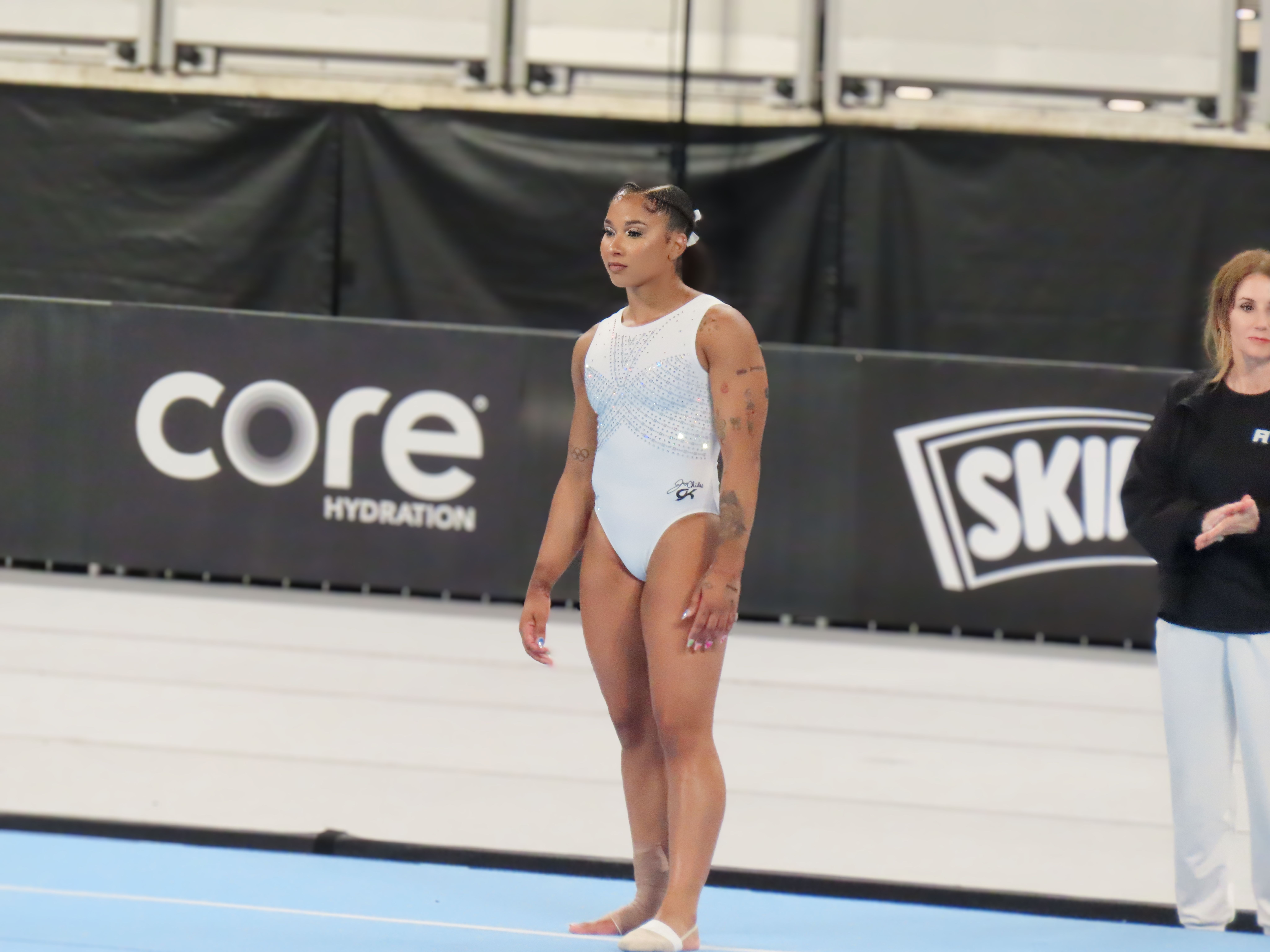 Gymnast in a white leotard standing on a competition floor exercise mat indoors.