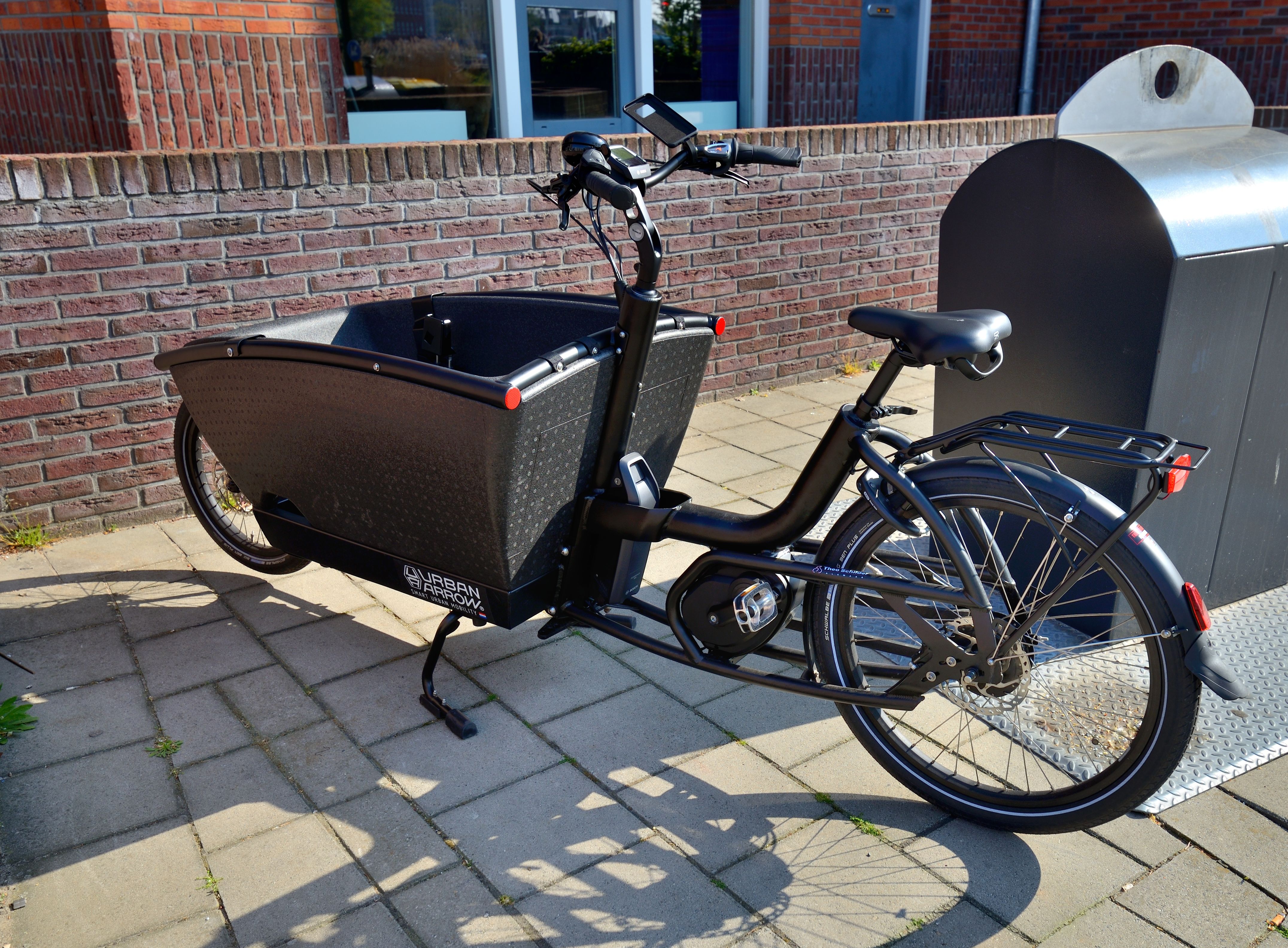 A black cargo e-bike with a large front box parked on a brick sidewalk.