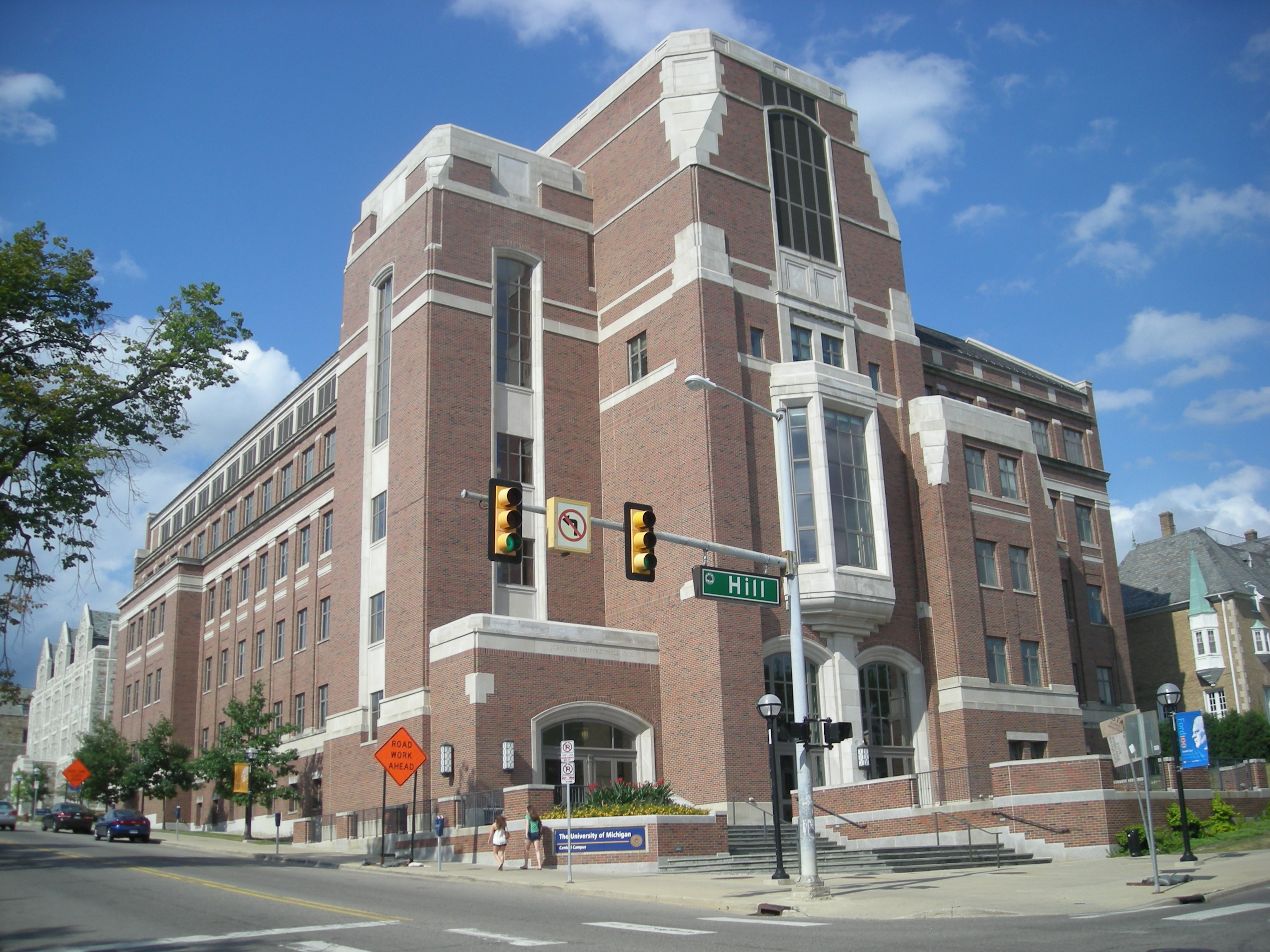 Red-brick multi-story academic building on a street corner under a partly cloudy sky.